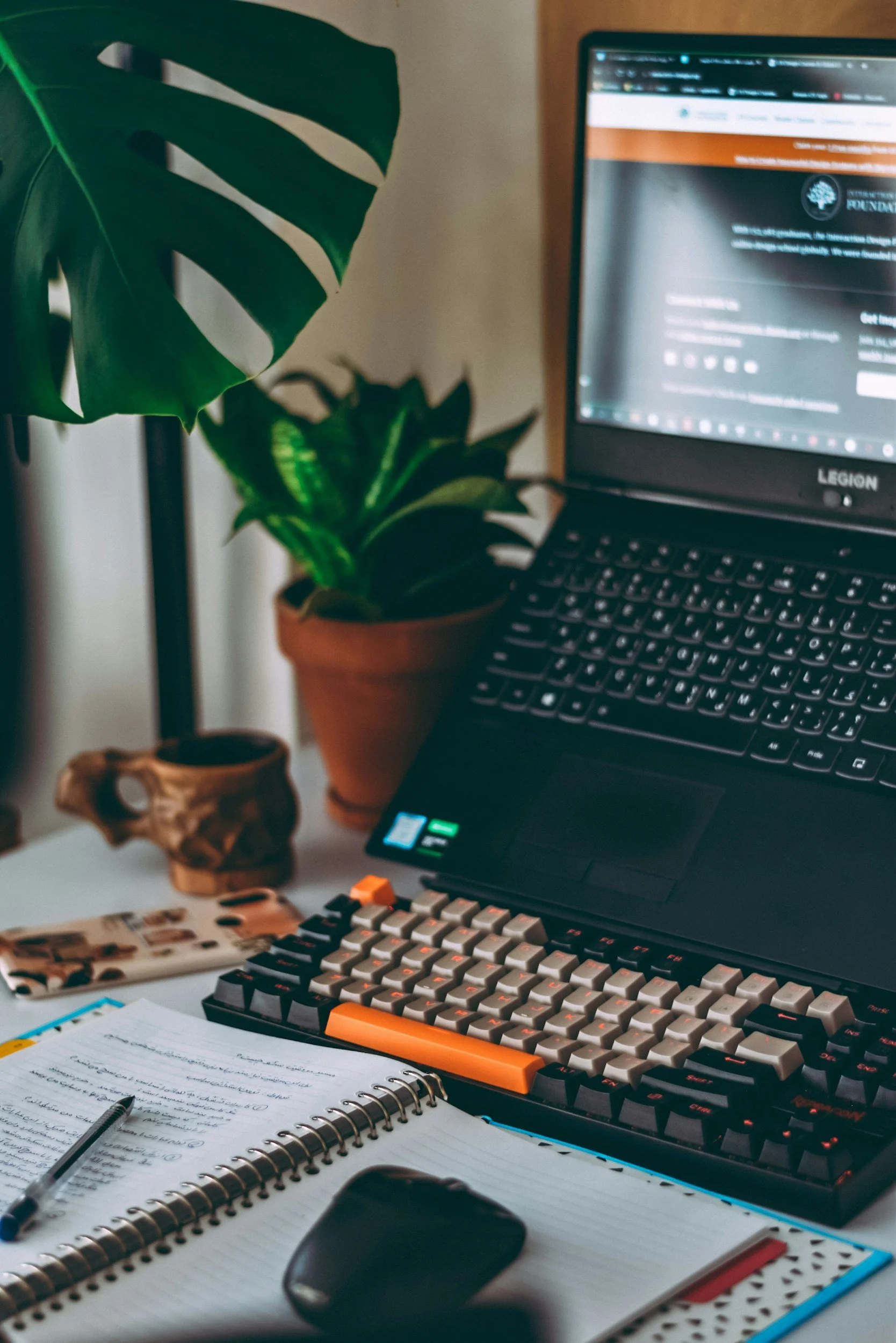 Home office desk with a computer laptop, mechanical keyboard, notebook, pen, and potted plants.