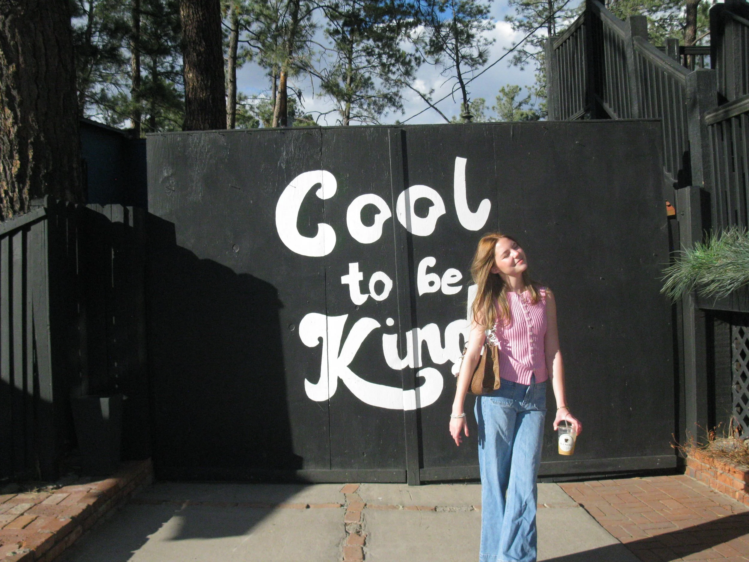A woman with shoulder-length hair, wearing a pink and white striped shirt and light blue jeans, standing outdoors in front of a black wooden fence with white lettering. She is holding a clear cup with a straw and appears to be enjoying the sunny day.