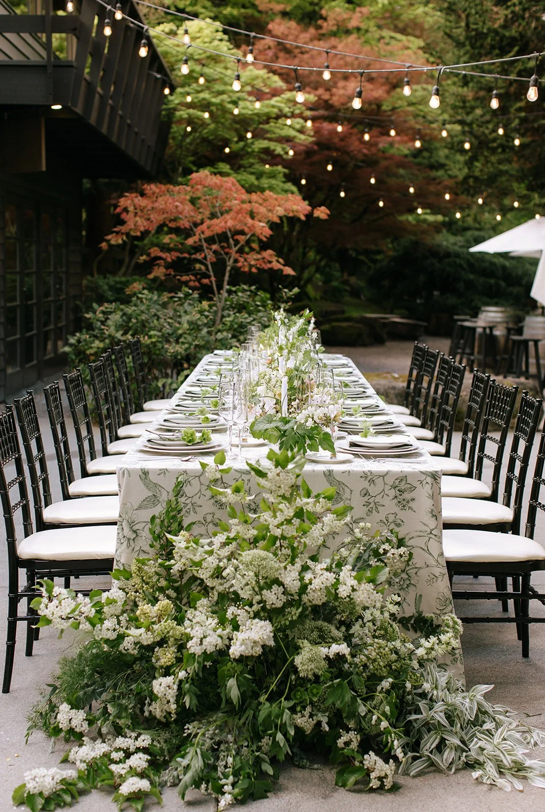 An elegantly set outdoor dining table covered with a floral-patterned tablecloth, decorated with lush greenery and white flowers. Surrounded by black chairs, the table is illuminated by string lights overhead. The background features a garden with colorful trees.
