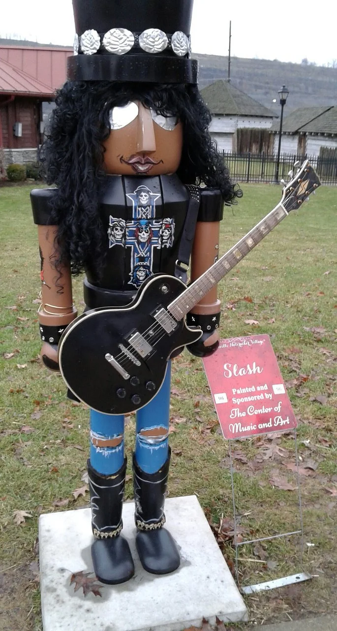 Decorative sculpture of a person with long black curly hair, wearing a black hat with silver decorations, holding a black electric guitar, with a patch on the shirt resembling a cross with skulls, and a red sign beside it that reads 'Slash, Painted and Sponsored by The Center of Music and Art'.