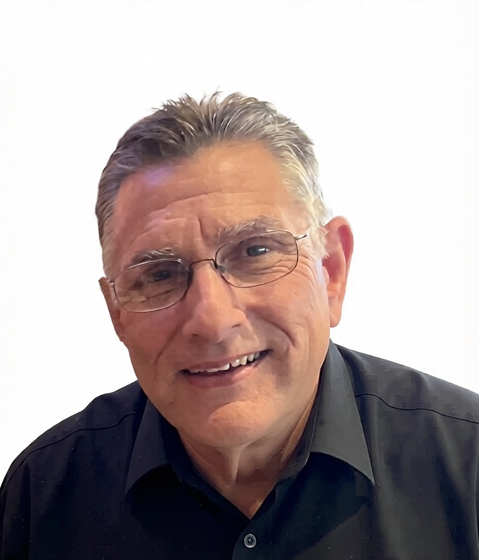 A man with gray hair, wearing glasses, a gray suit, a light green shirt, and a colorful patterned tie, smiling at the camera against a green background.