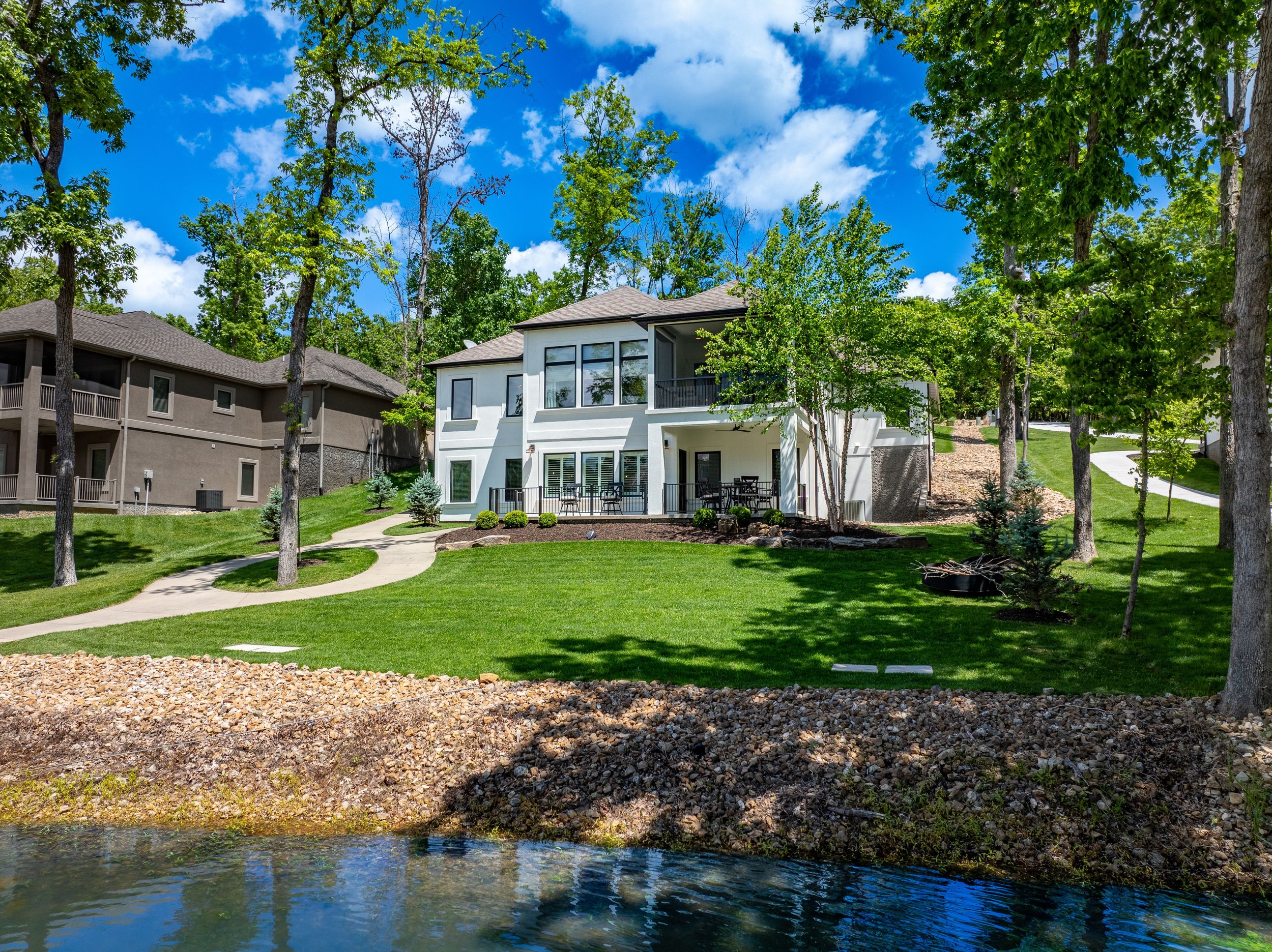 Modern two-story house with large windows, surrounded by trees and a well-maintained lawn, located near a small body of water under a clear blue sky.