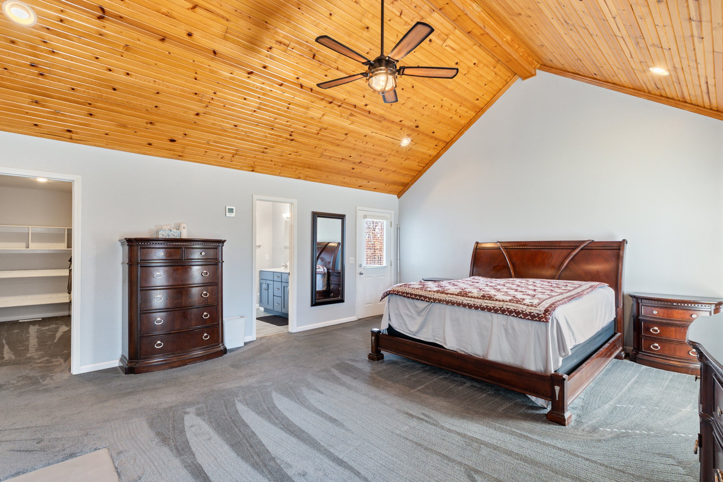 Spacious bedroom with wooden ceiling, ceiling fan, large bed with wooden frame, dresser, nightstand, carpeted floor, and visible doorways to closet and bathroom.