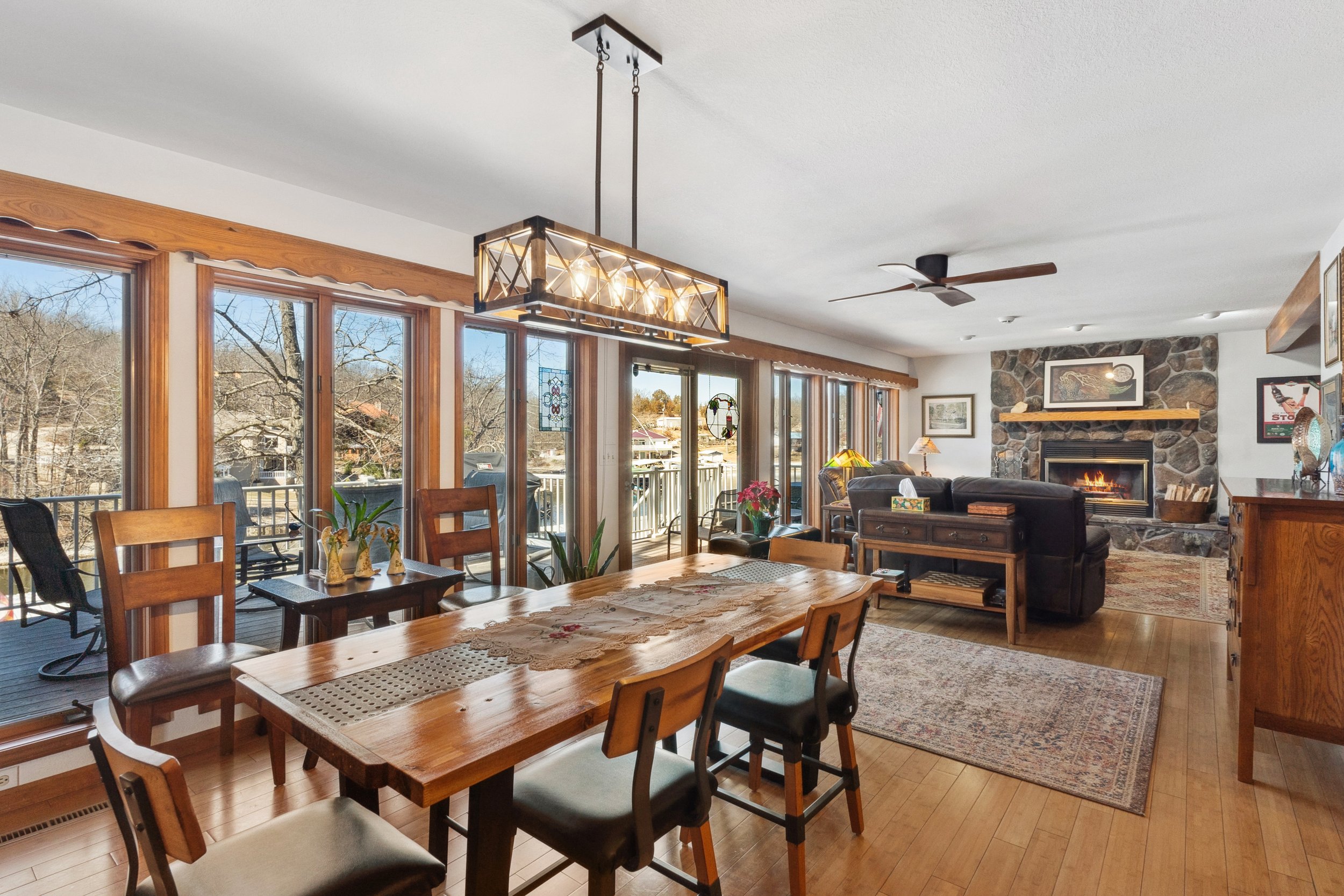 Open-concept dining and living room with a wooden table, chairs, and a large chandelier. The room features a stone fireplace, brown sofas, and hardwood flooring. Large windows with a view of the outdoors run along one side, letting in natural light.