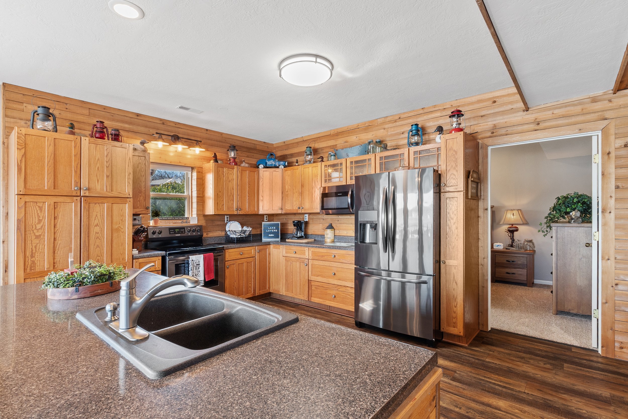 Rustic kitchen with wooden cabinets, stainless steel appliances, and a granite countertop. Decor includes vintage lanterns and plants. View into an adjoining room with wooden furniture and decor.