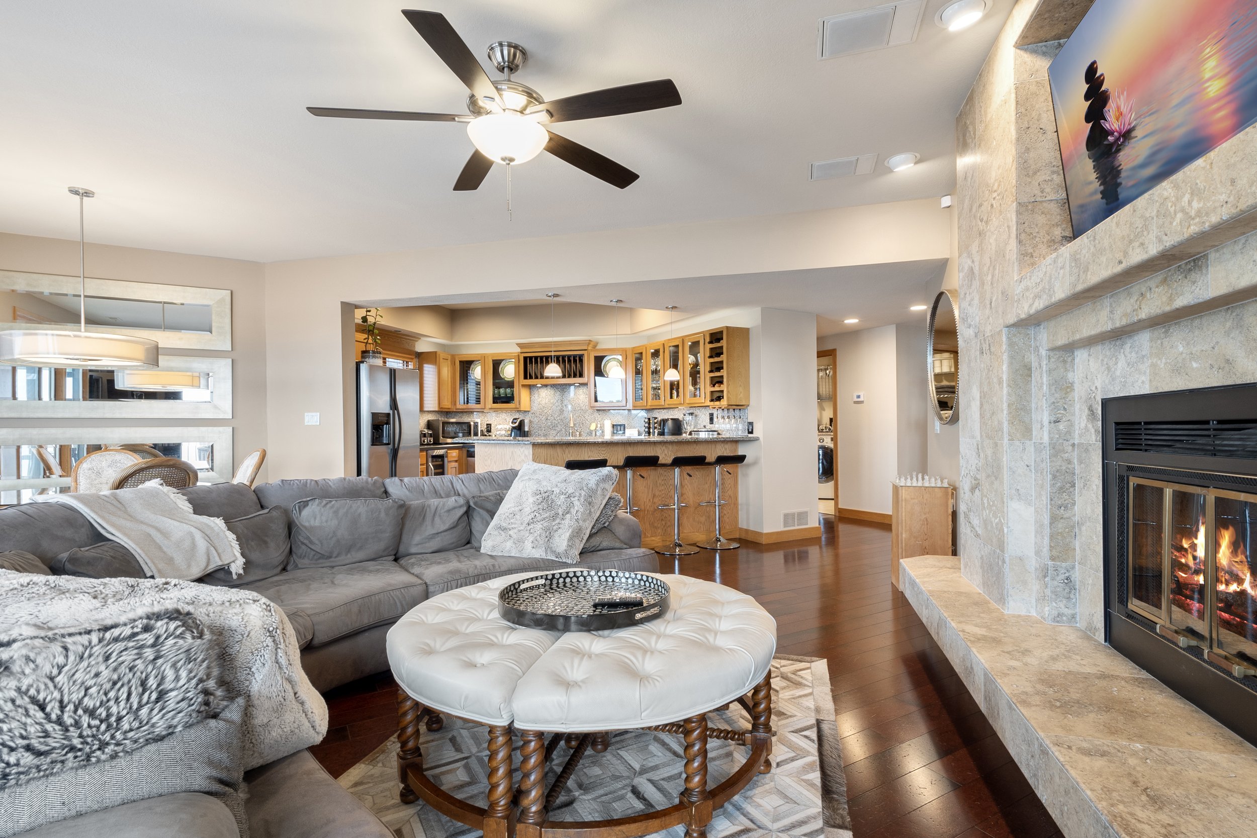 Modern living room with a gray sectional sofa, a round white ottoman, a fireplace, hardwood floors, and an open kitchen. The kitchen features wooden cabinets and a breakfast bar with black stools. A ceiling fan and contemporary lighting are present.