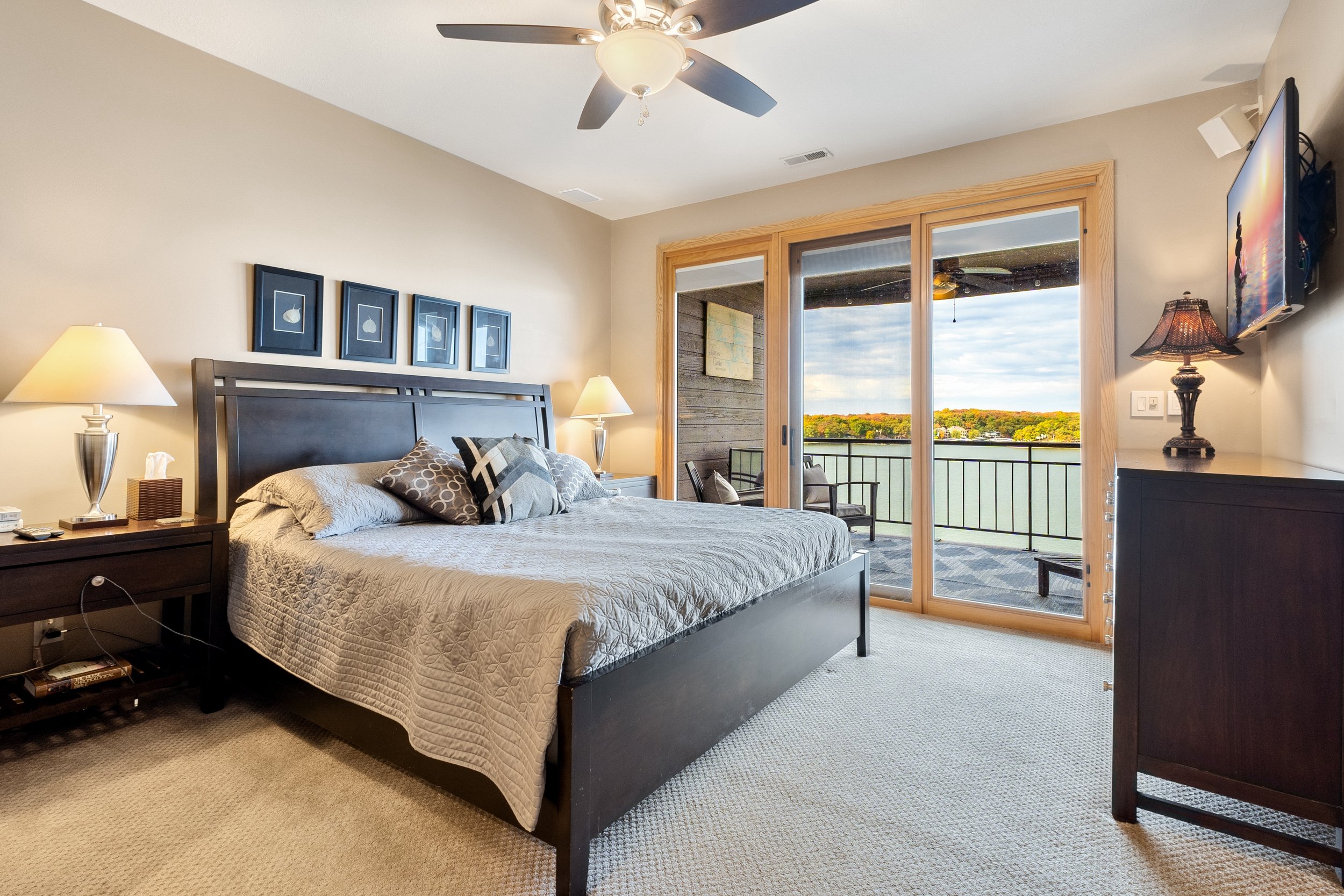 Modern bedroom with king-sized bed, decorative pillows, two bedside lamps, decorative wall art, ceiling fan, and glass sliding doors leading to a balcony with outdoor seating and a scenic lake view.