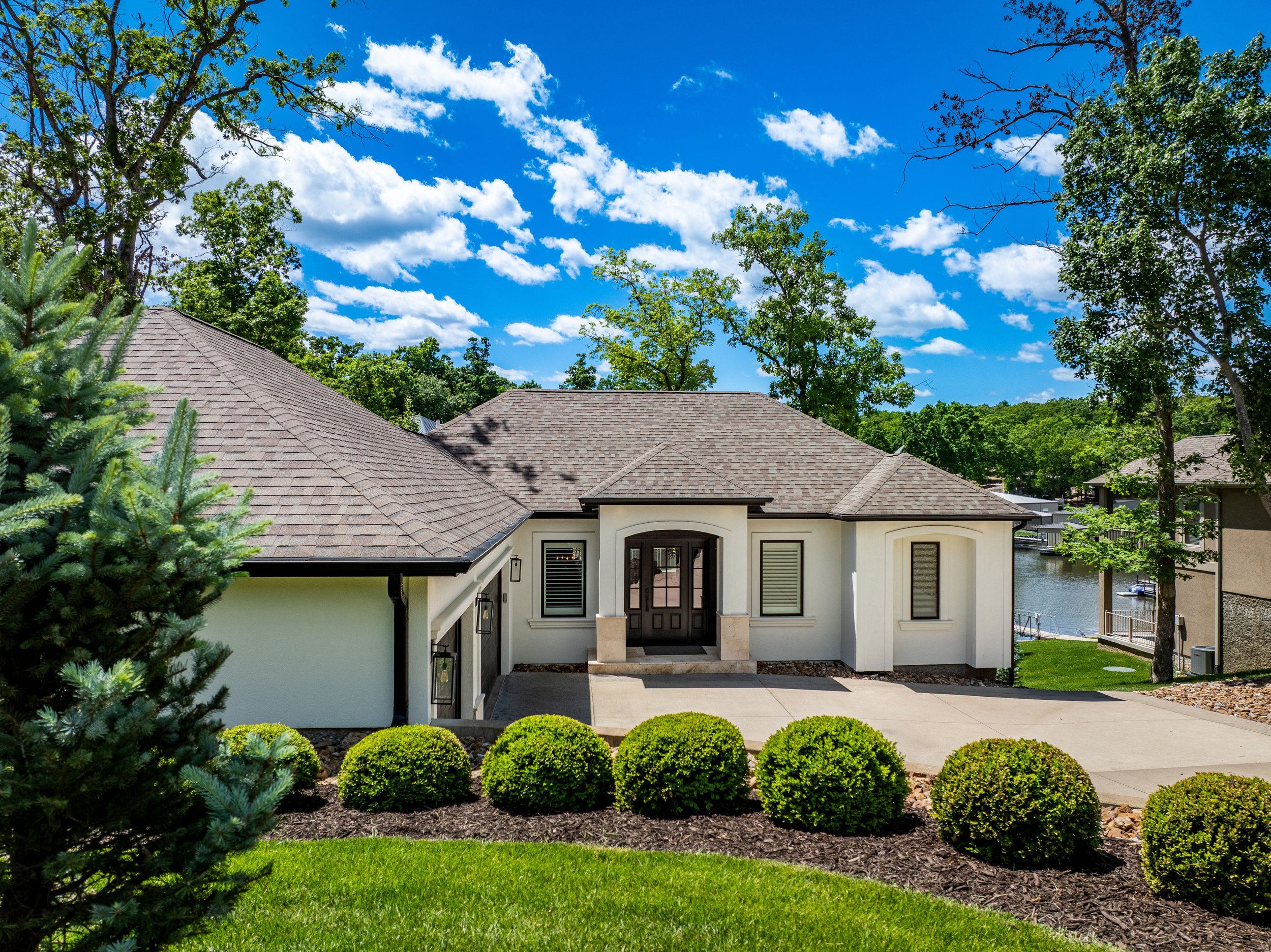 A modern single-story house with a light-colored exterior, surrounded by well-maintained landscaping, including bushes and grass. The roof is made of shingles, and the entrance features double doors. Trees and a clear blue sky are in the background.