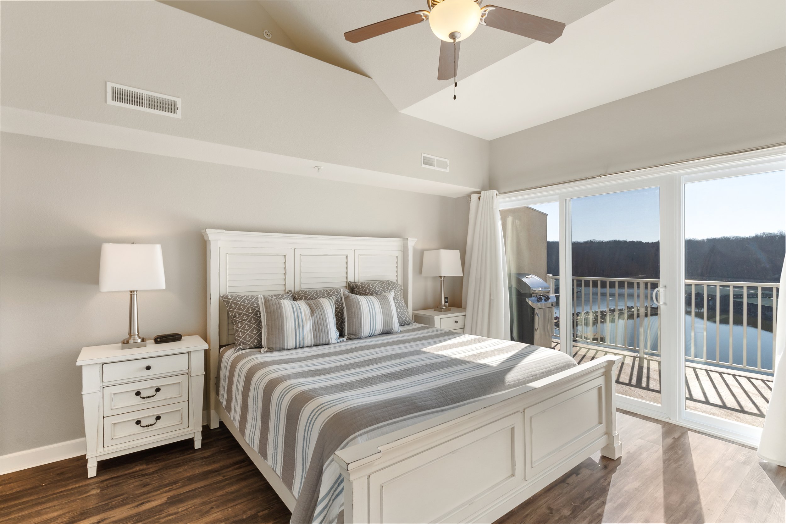 Bright bedroom with striped bedspread, bedside tables with lamps, ceiling fan, sliding glass doors leading to a balcony with lake view.