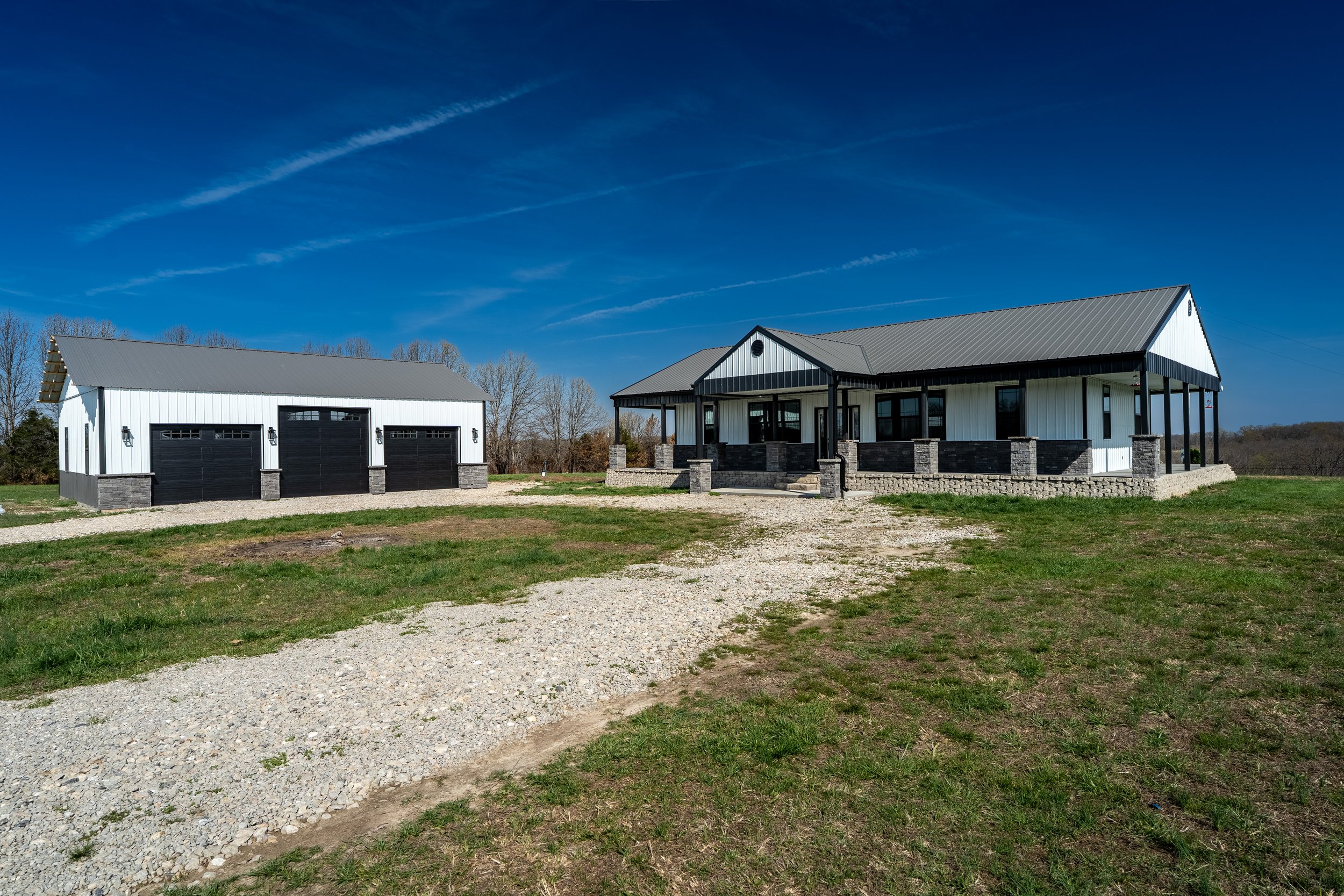 Modern farmhouse with a metal roof and detached three-car garage in a rural setting under a clear blue sky.