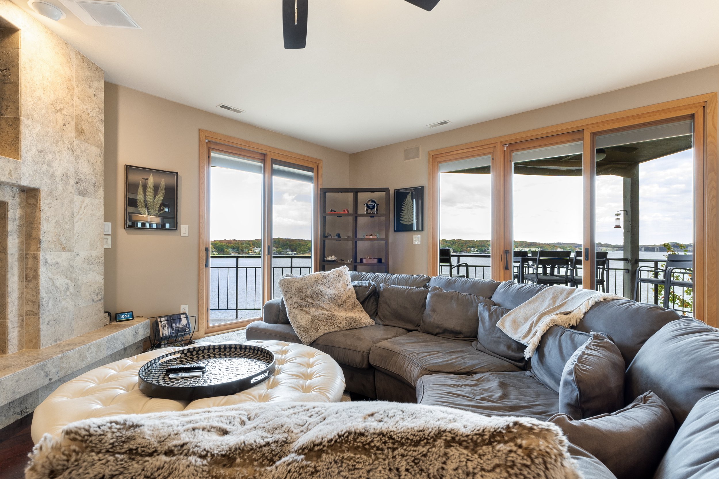 Modern living room with a large gray sectional sofa, fluffy pillows, and a stylish ottoman. Room features wooden framed sliding glass doors opening to a scenic balcony view of a lake. A stone fireplace and open shelving are visible, creating a cozy a