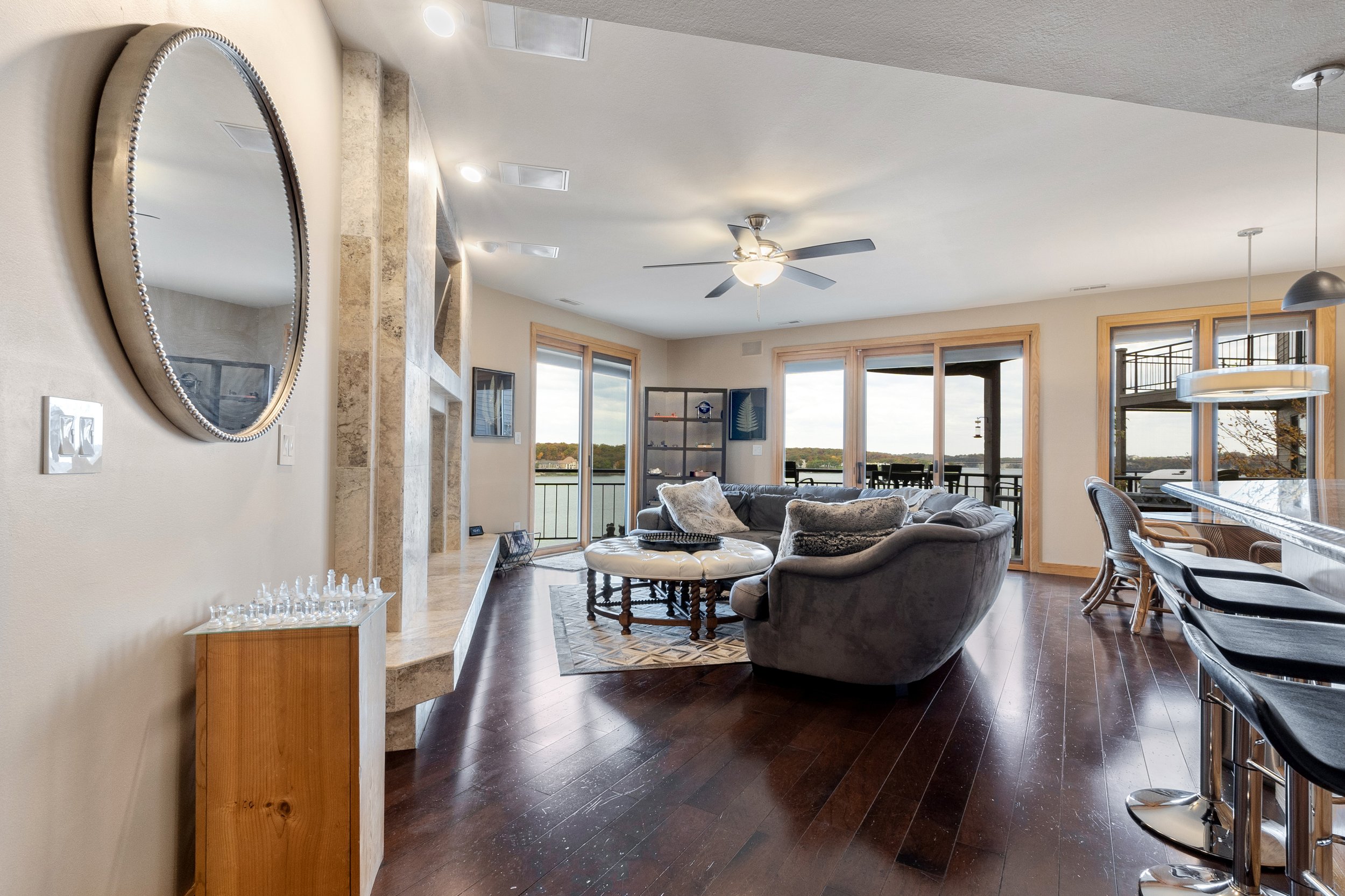 Stylish living room with large windows, gray sofas, round coffee table, ceiling fan, bar stools, and a wall mirror. Wooden floor and view of outdoor patio.
