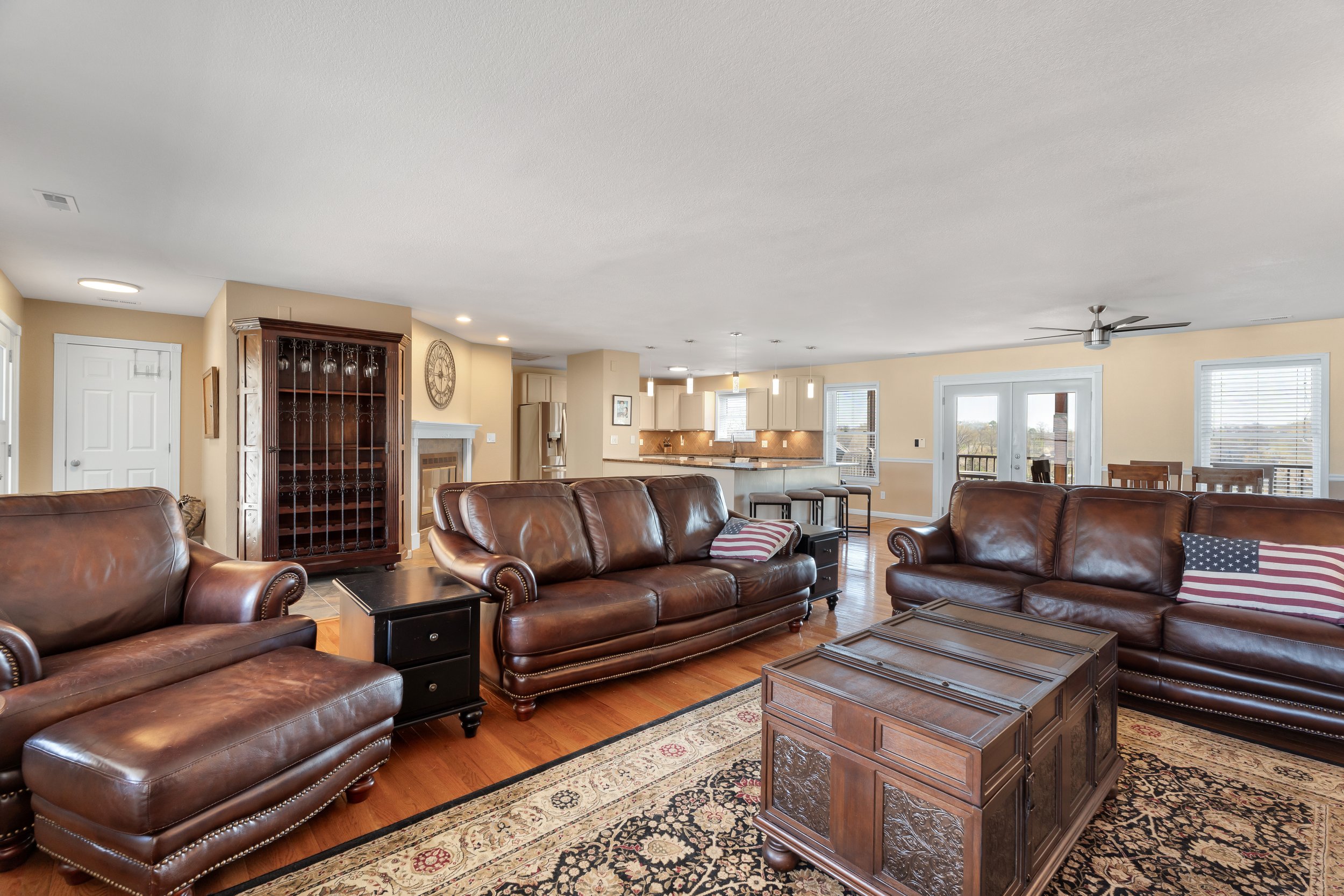 Spacious living room with brown leather sofas, a patterned area rug, wooden coffee table, and open kitchen in the background. An American flag pillow is on one sofa and a ceiling fan is overhead. The room is well-lit with large windows and hanging li