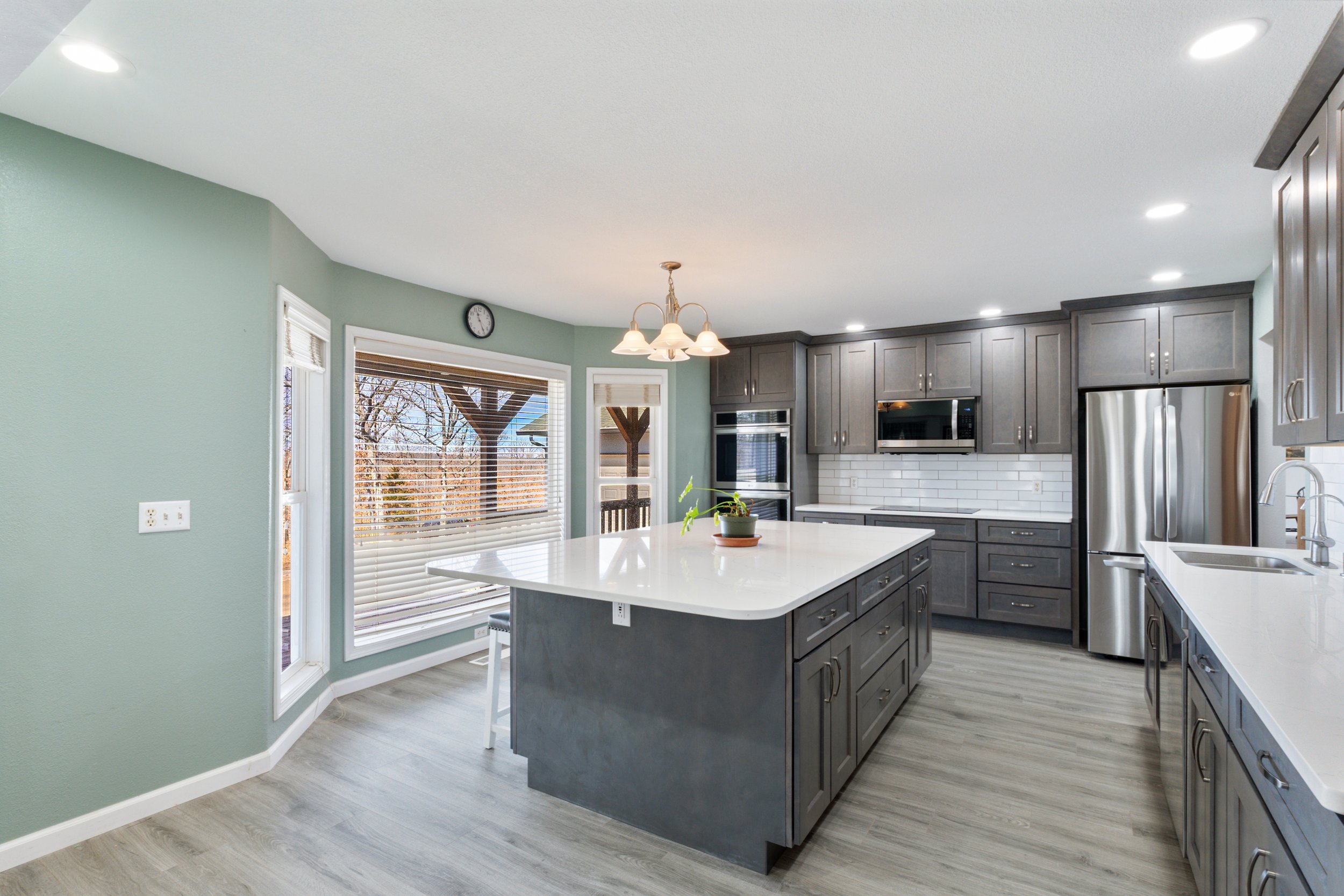 Modern kitchen with gray cabinets, white countertops, and a center island. Stainless steel appliances include a refrigerator, microwave, and double ovens. Large windows with blinds offer natural light. A clock and pendant lights are present.