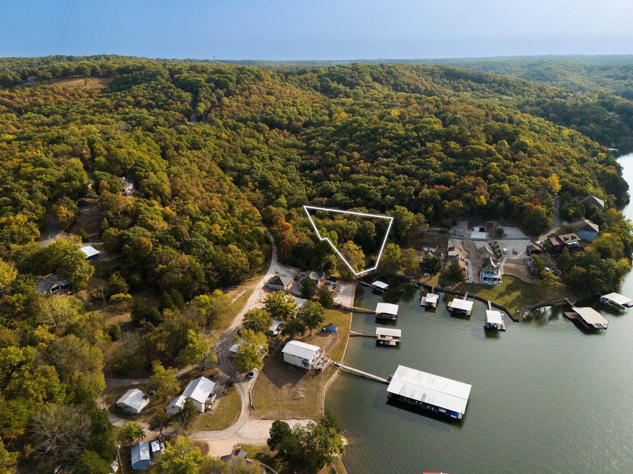 Aerial view of a wooded lakeside area with scattered buildings and docks. The area features a mix of dense trees and open spaces, with a body of water visible and several boat docks along the shoreline. A highlighted section indicates a specific plot