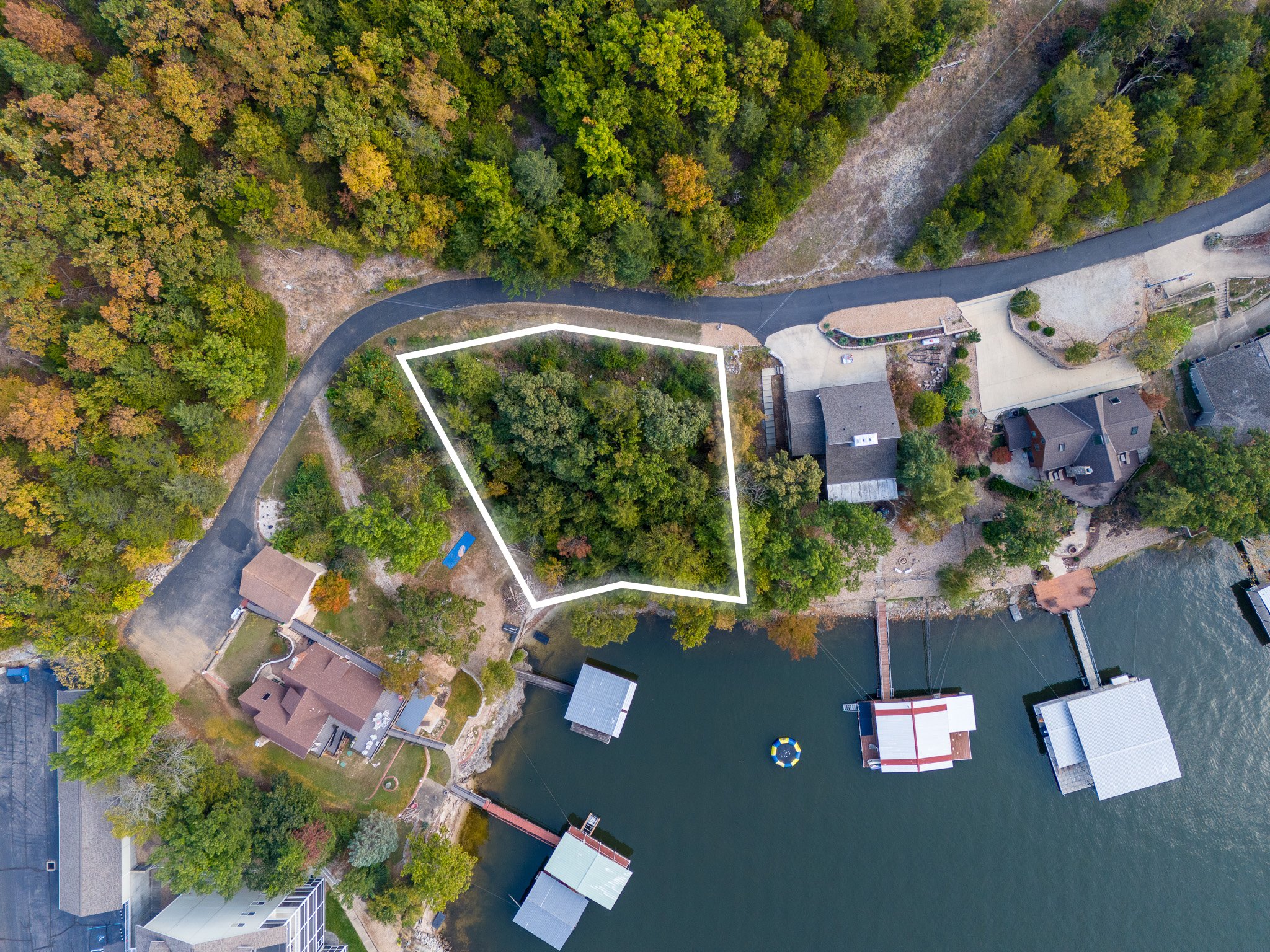 Aerial view of a residential area by a lake, with houses, trees, and docks. A plot of land is outlined in white.