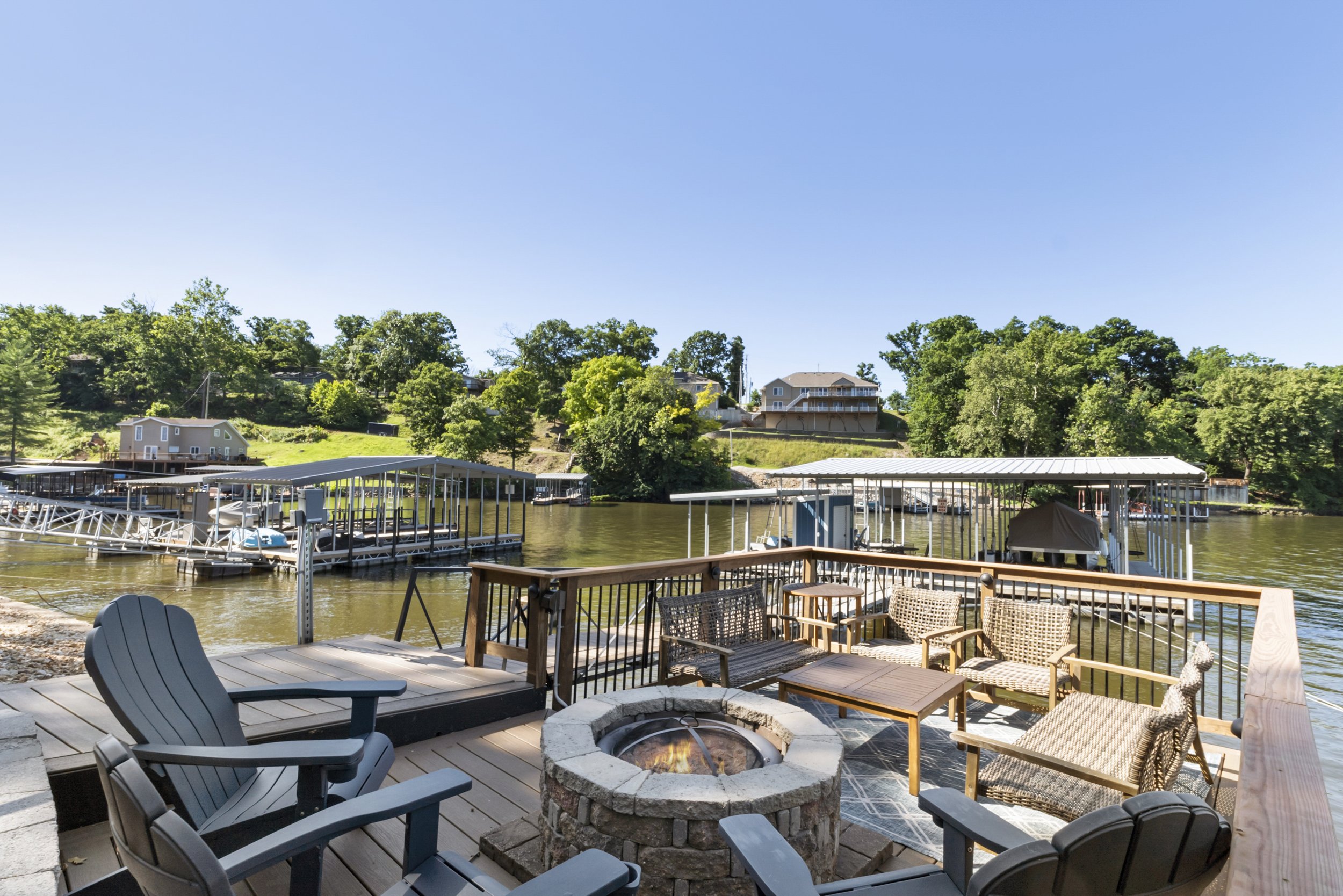 Lakefront patio with chairs and fire pit overlooking docks and water, surrounded by trees and houses in the background.