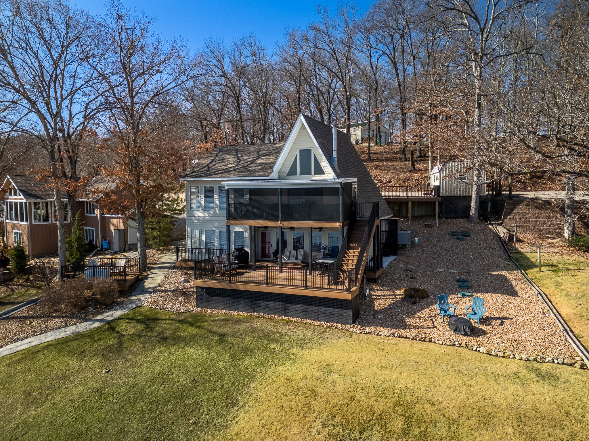A-frame house with large deck, surrounded by leafless trees, on a sloped yard with gravel patio area featuring chairs and fire pit; another house is visible nearby.