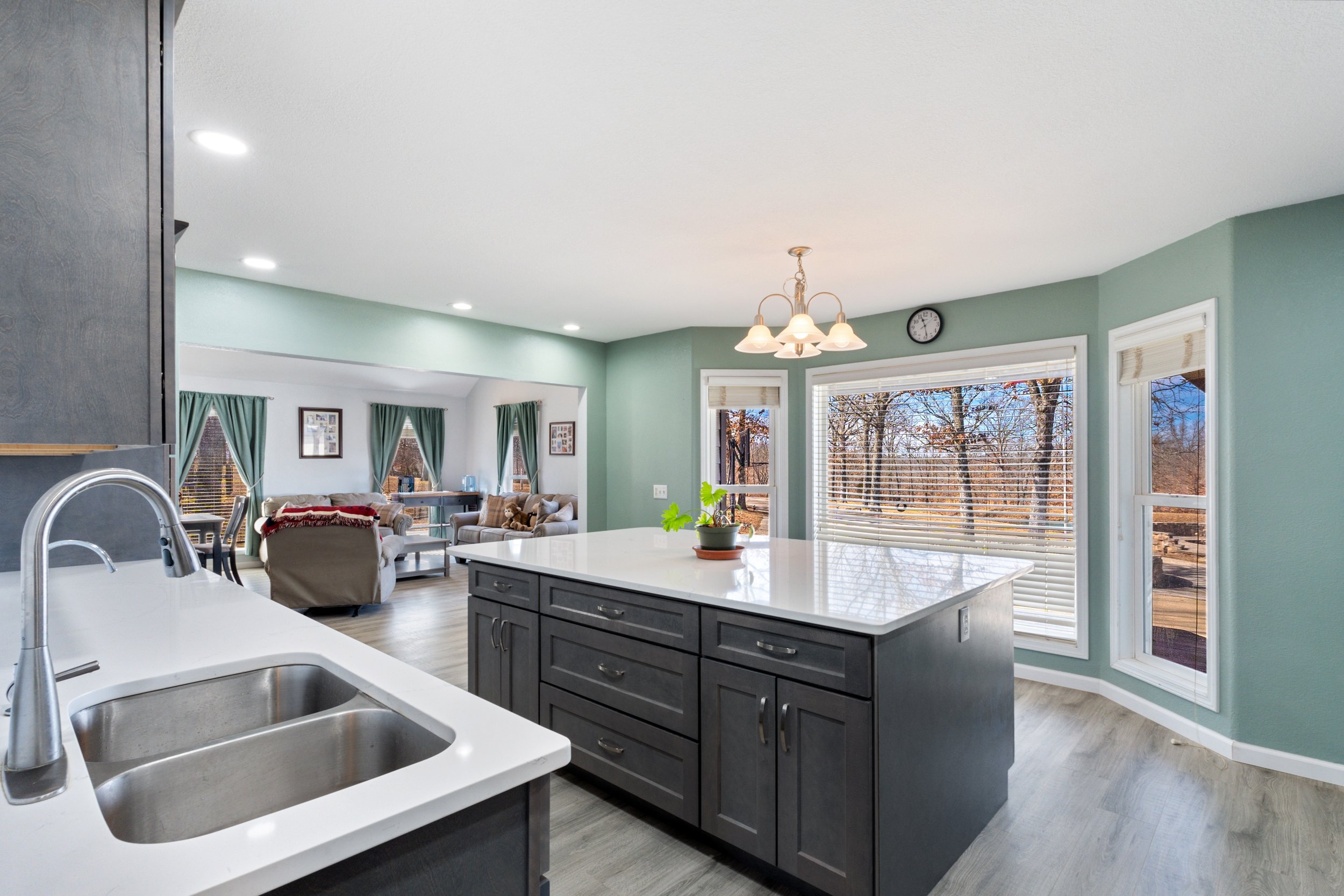 Modern kitchen interior with a large island, double sink, and a view of a living area with green curtains. The room features light green walls, recessed lighting, a chandelier, and large windows offering natural light and an outdoor view.