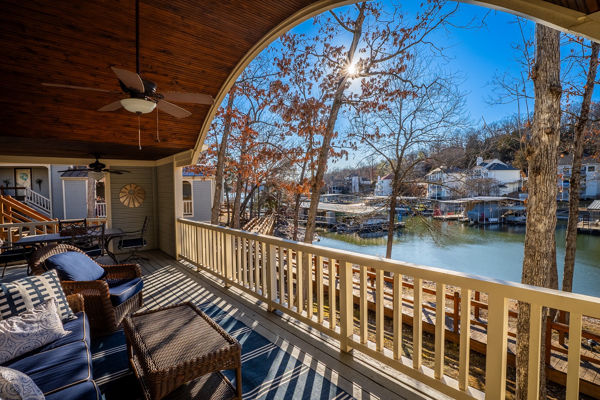 Covered waterfront patio with wicker furniture, overlooking a lake with trees, docks, and houses in the background, under blue sky.