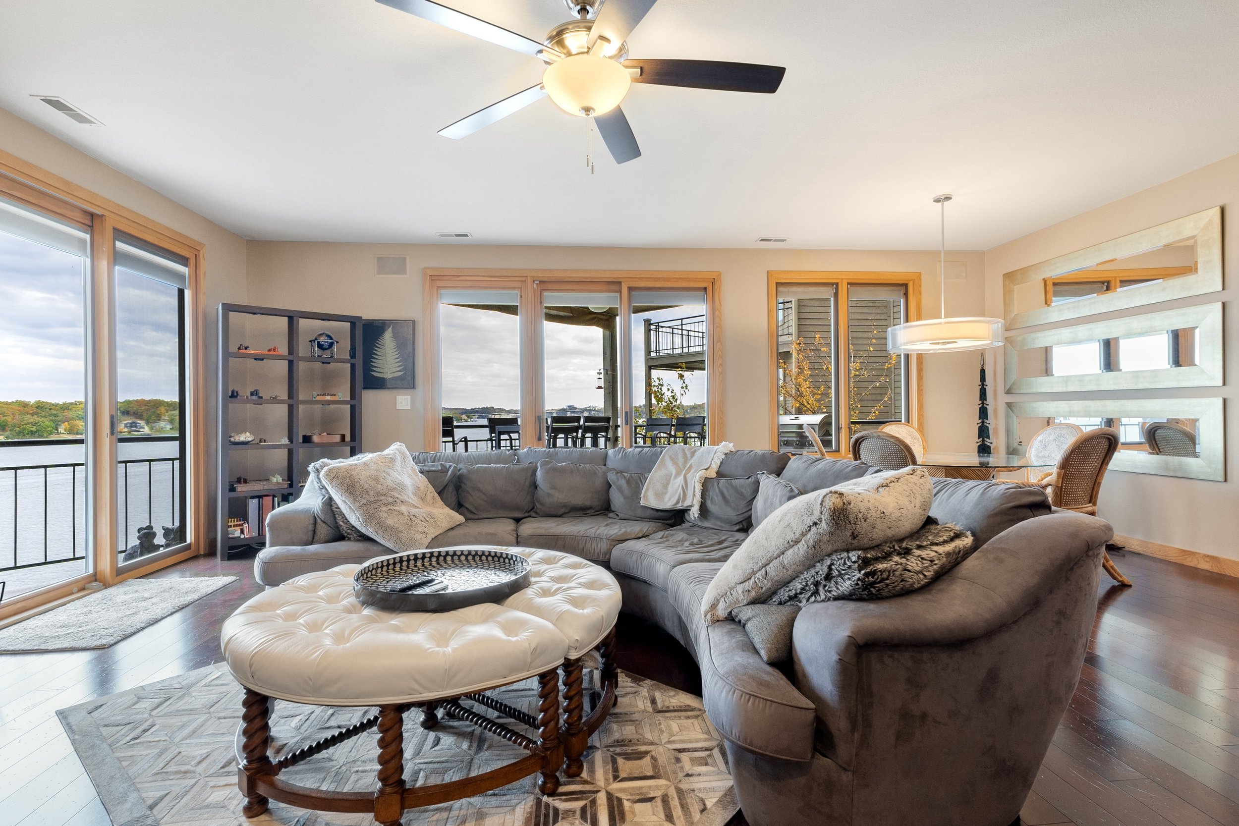 Modern living room with gray sectional sofa, round ottoman, ceiling fan, and large windows overlooking water view.