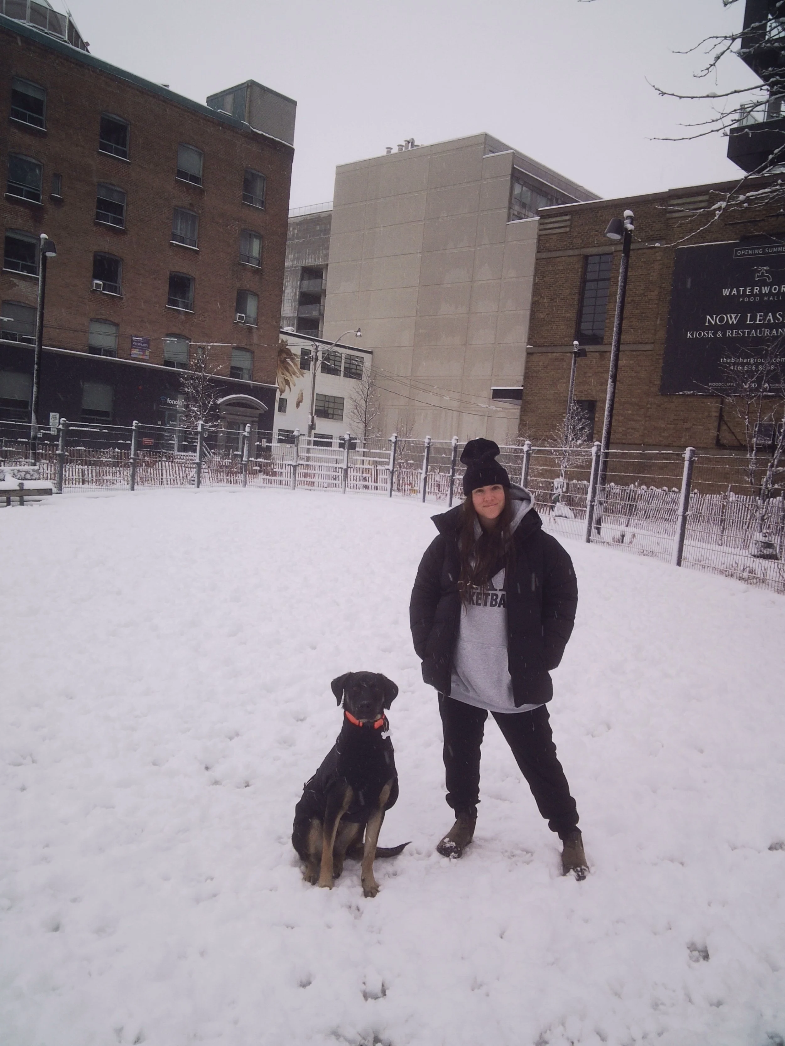 A woman and a dog standing in a snow-covered park with buildings in the background.