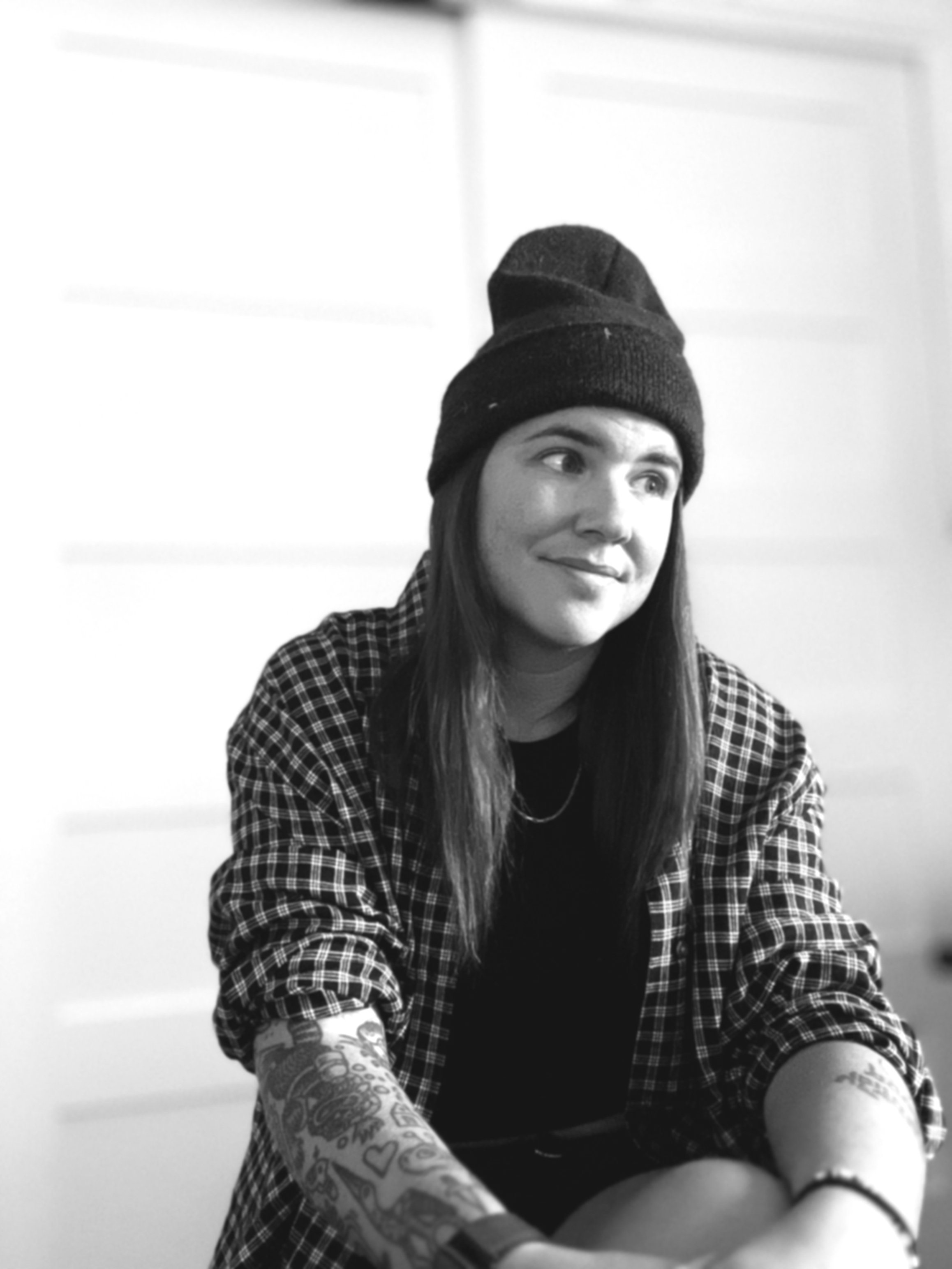 A young woman with long hair wearing a beanie, striped shirt, and tattooed arms, sitting indoors with a slight smile.