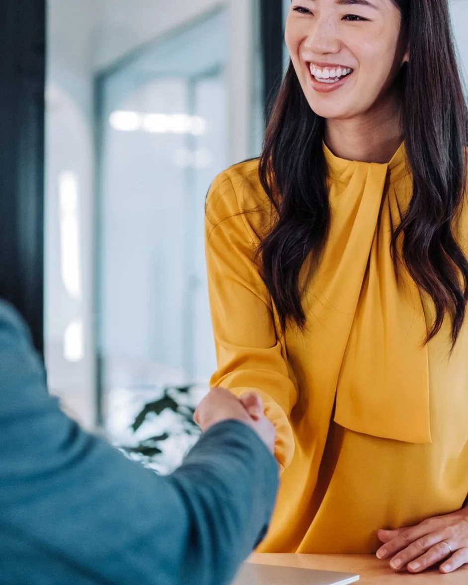 A woman in a yellow blouse smiles while shaking hands with another person in an office setting.