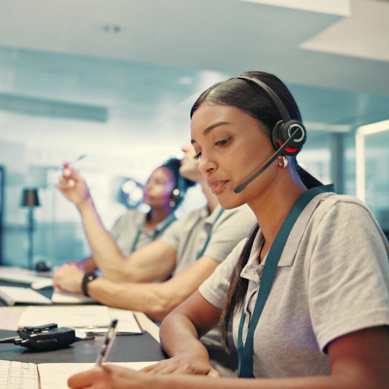 Woman wearing a headset takes notes at a desk in a call center with colleagues working in the background.
