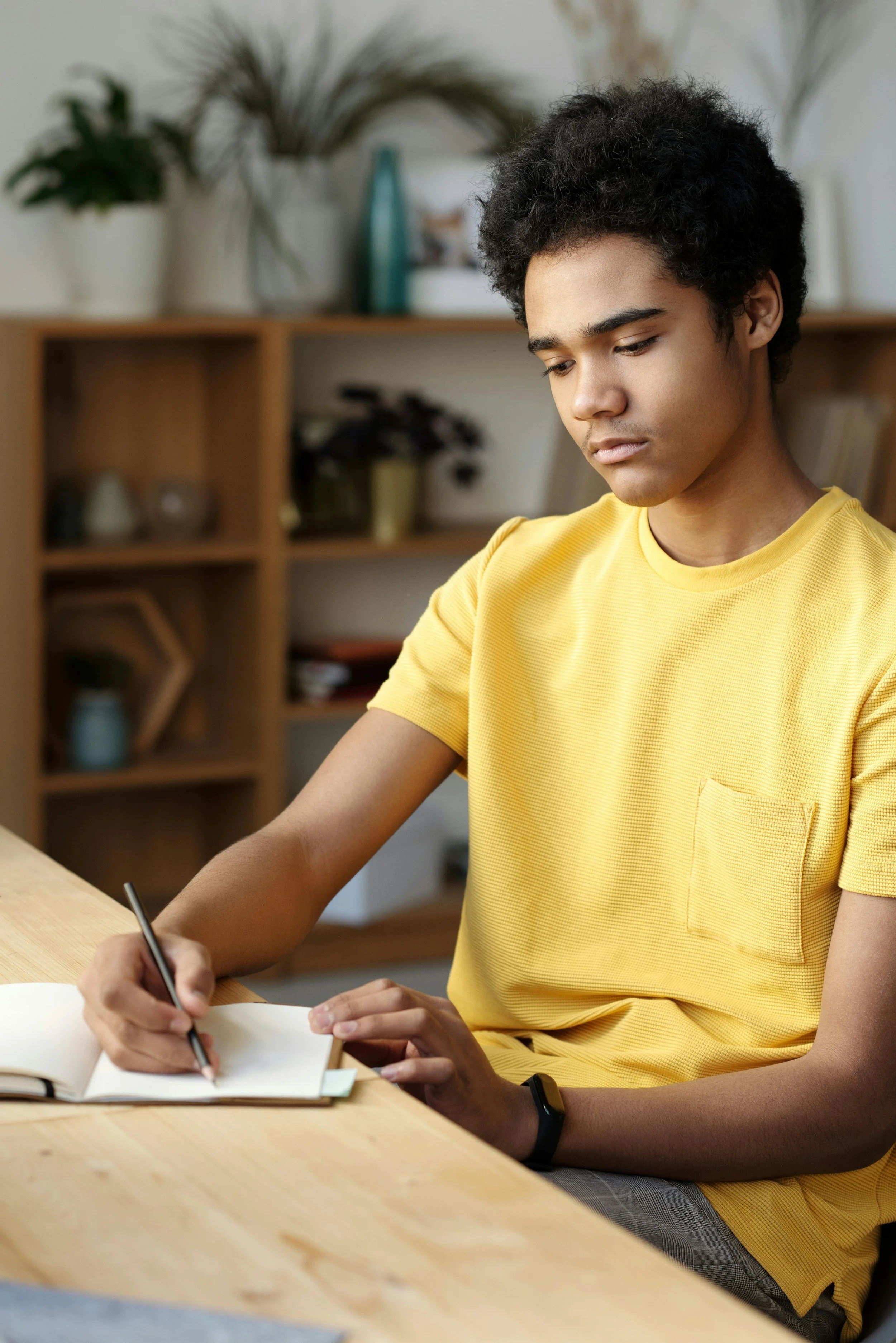 A college student is in a yellow shirt and sits at a table, writing in a notebook with a pencil. Shelves and plants are in the background.