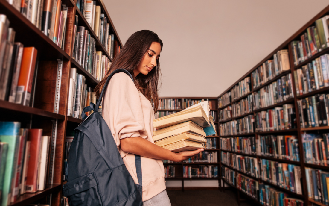 Woman in a library holding a stack of books, wearing a backpack, surrounded by bookshelves filled with various titles.