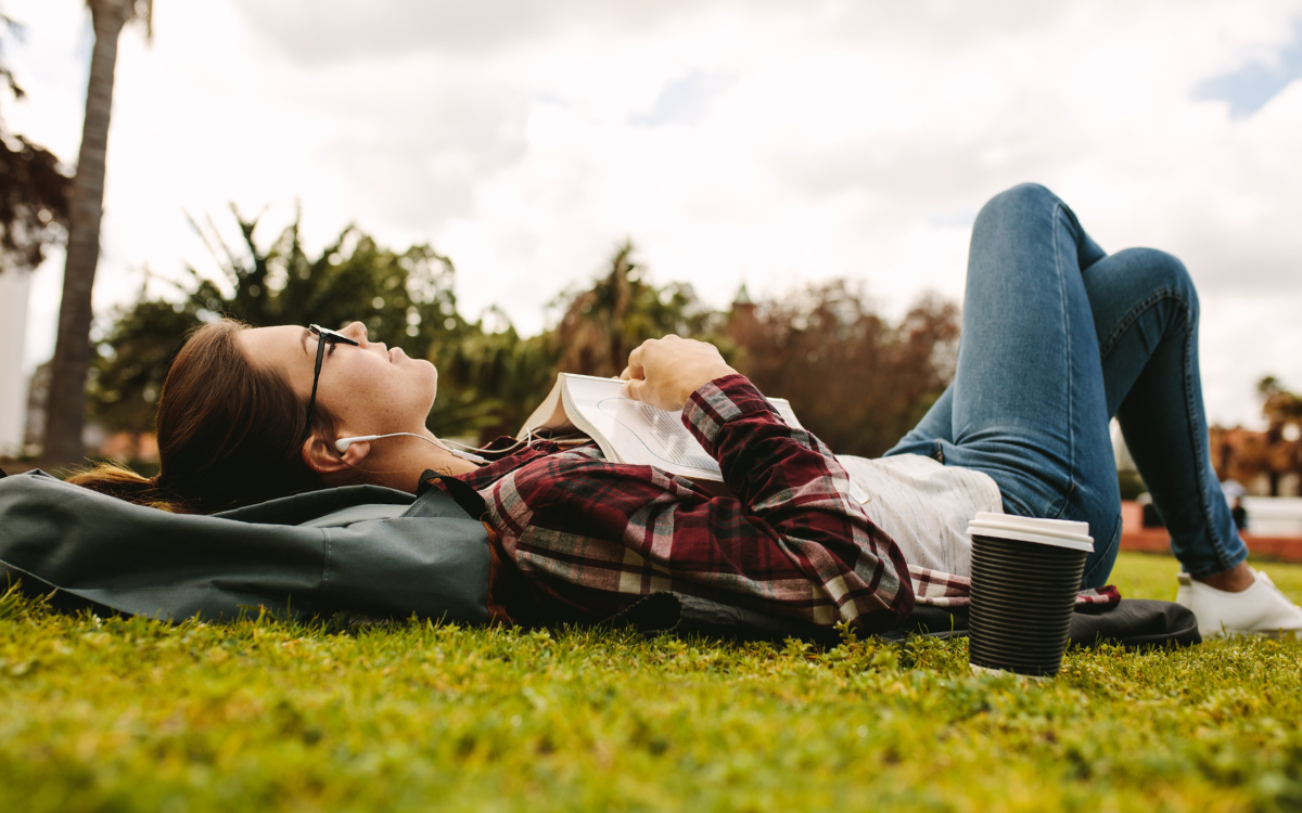 Person lying on grass with headphones and an open book, wearing a flannel shirt and jeans, next to a cup of coffee.