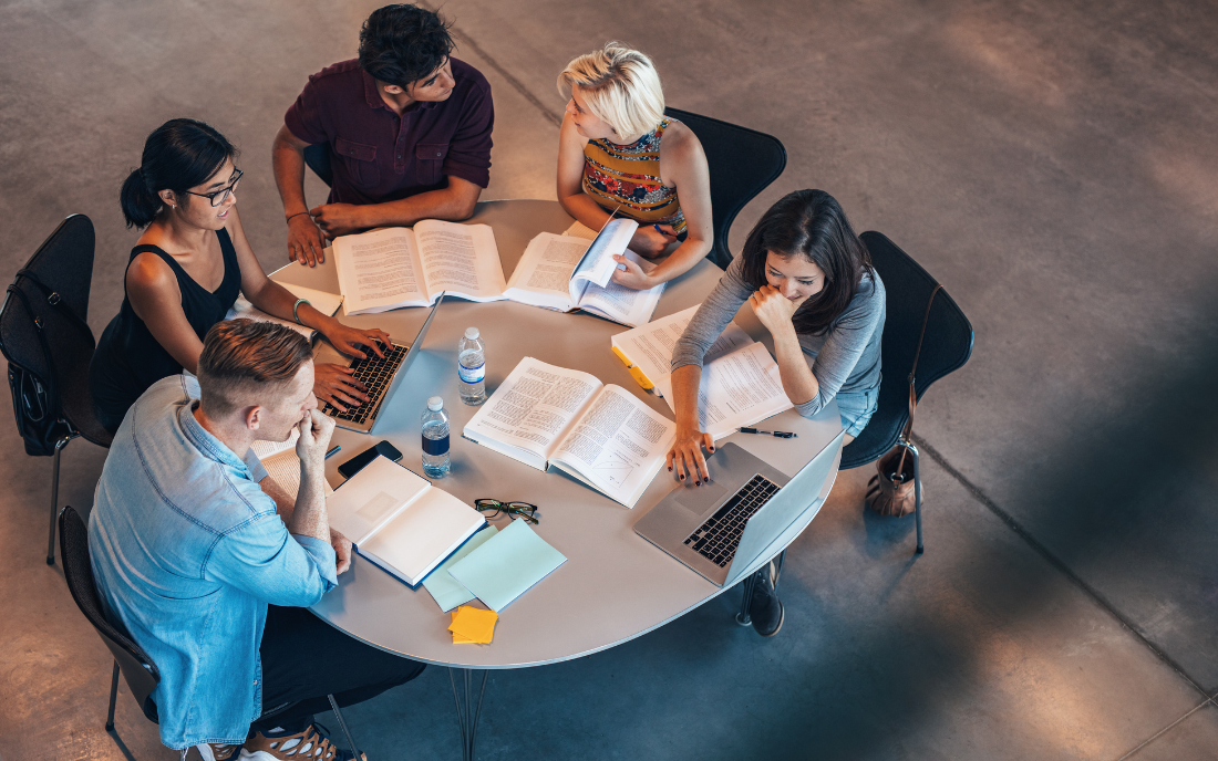 Five people sitting around a round table with laptops and open books, engaged in discussion or study in a spacious room.