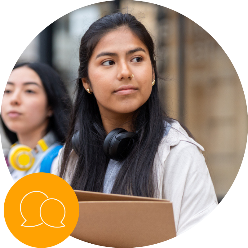Young college student with headphones around her neck holds a box outdoors, with another person blurred in the background.