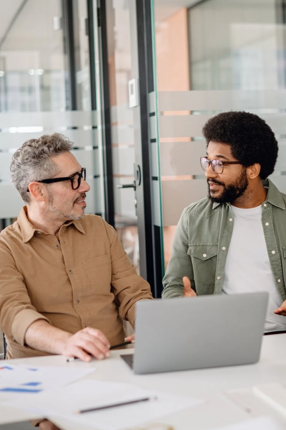 Two men sit in a workplace with a laptop, engaged in conversation in a modern office setting.