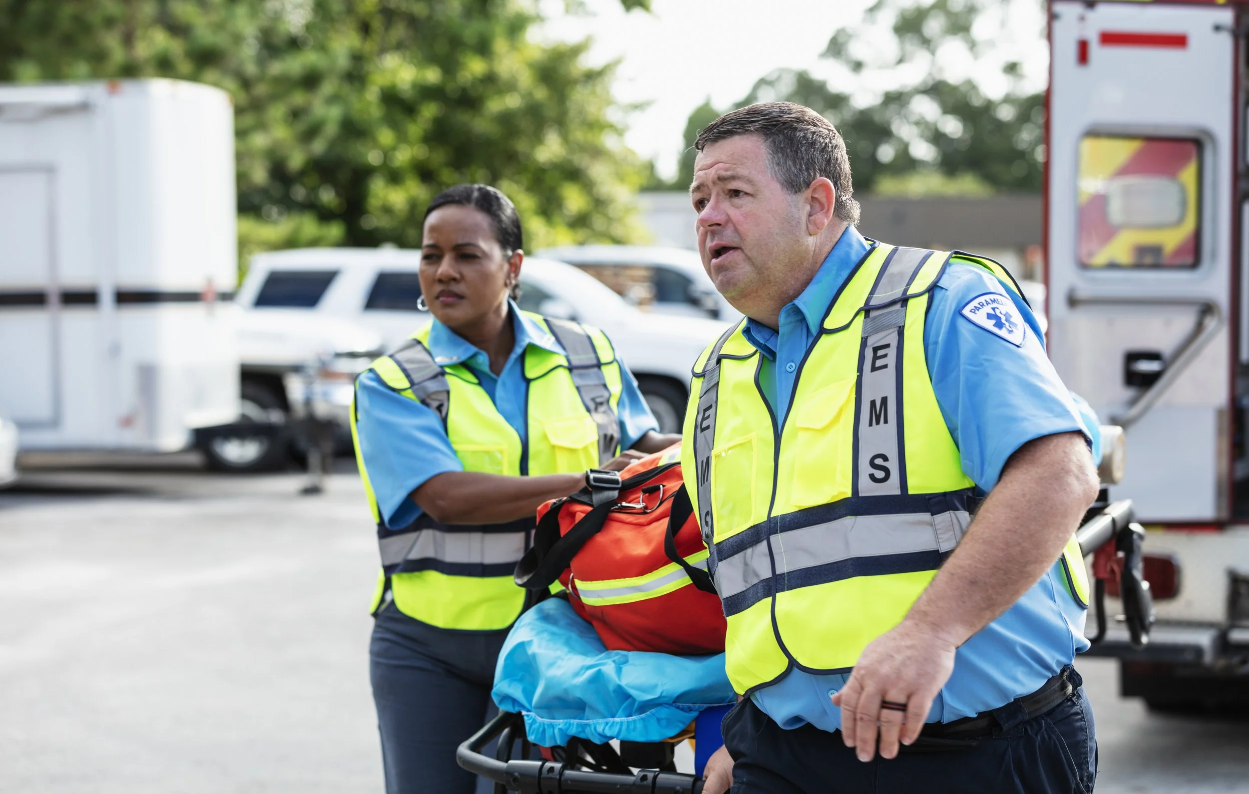 Two EMS workers in uniforms and safety vests transport medical equipment on a stretcher outdoors.