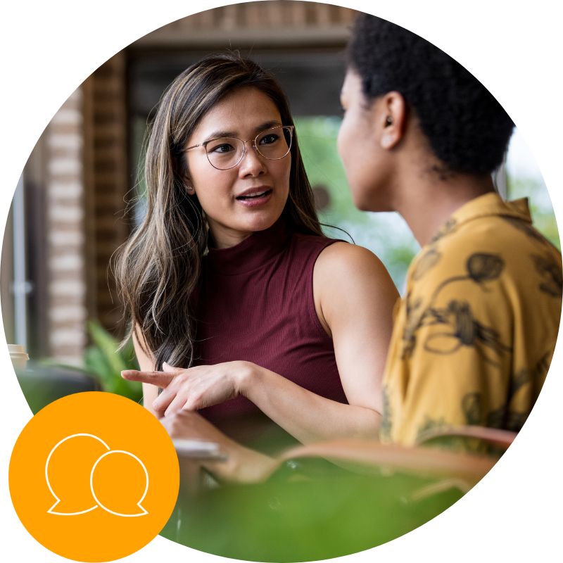 Two people sit indoors having a focused conversation; one gestures while speaking to the other.