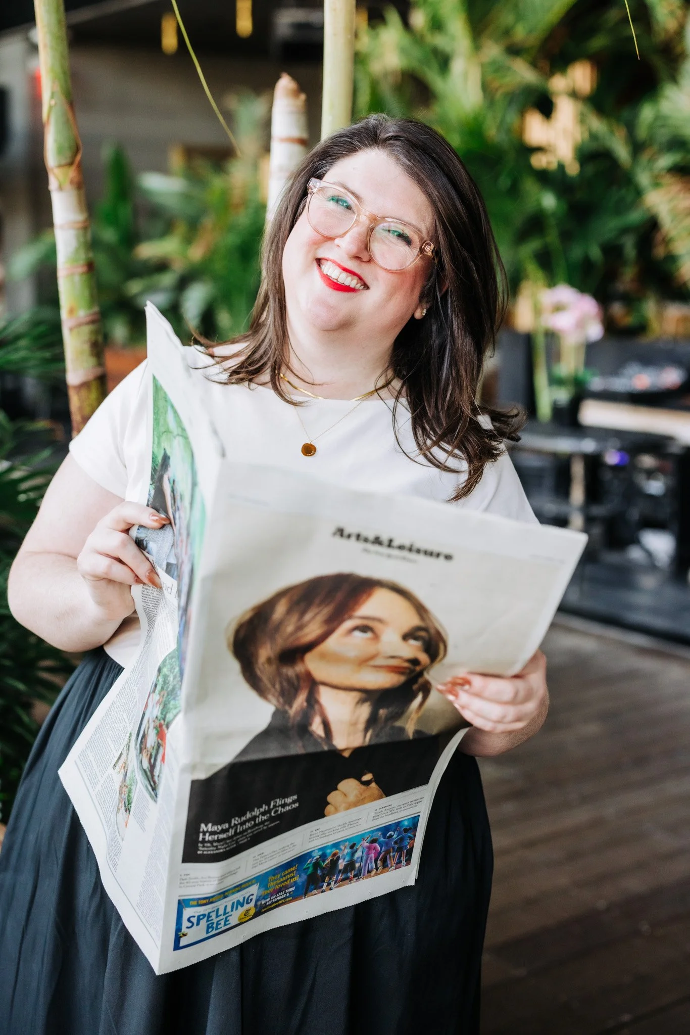 Woman with glasses and red lipstick holding a newspaper titled 'Art & Leisure', smiling, in an outdoor setting with plants in the background.