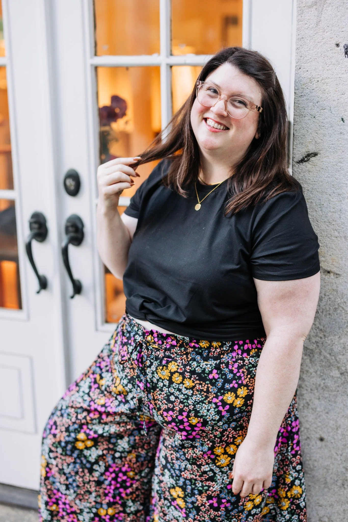 A smiling woman with glasses and long brown hair, wearing a black T-shirt and floral pants, standing outdoors near a stone wall and a window with orange lighting inside.