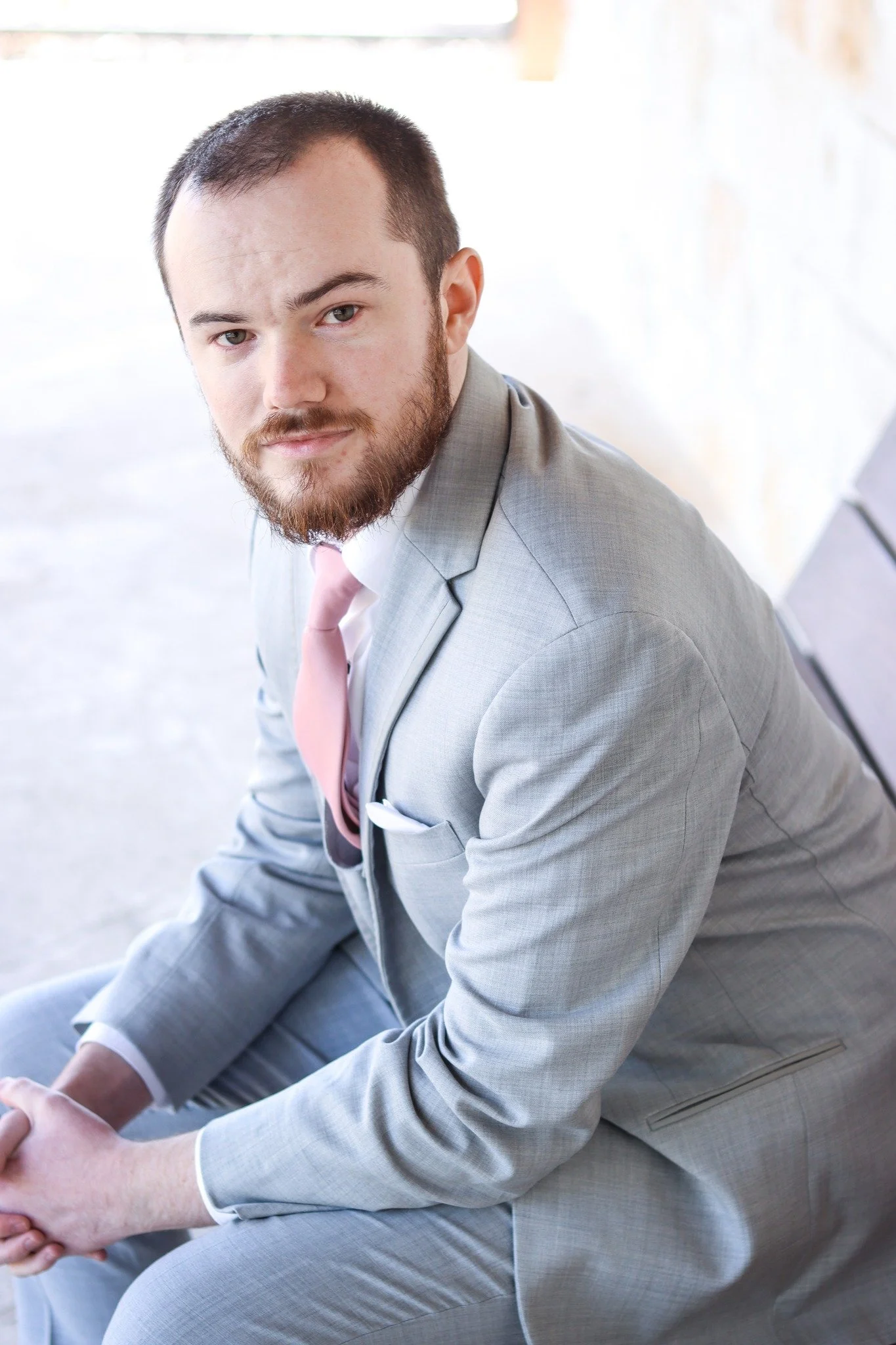 A man with a beard and short hair, wearing a light gray suit and a pink tie, sitting outdoors with a bright background.
