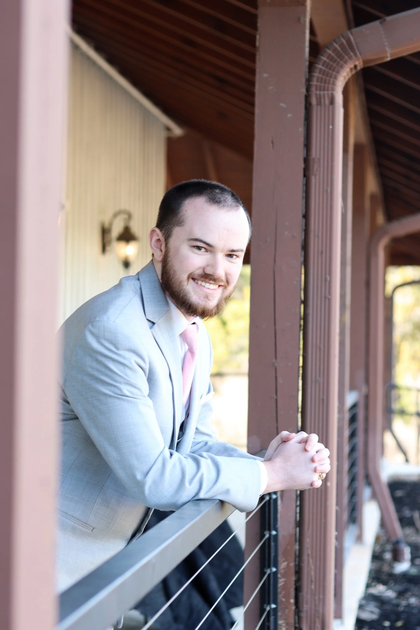 A young man wearing a light gray suit, white shirt, and pink tie, smiling and leaning on a railing on a porch with a wooden roof and exterior lighting.