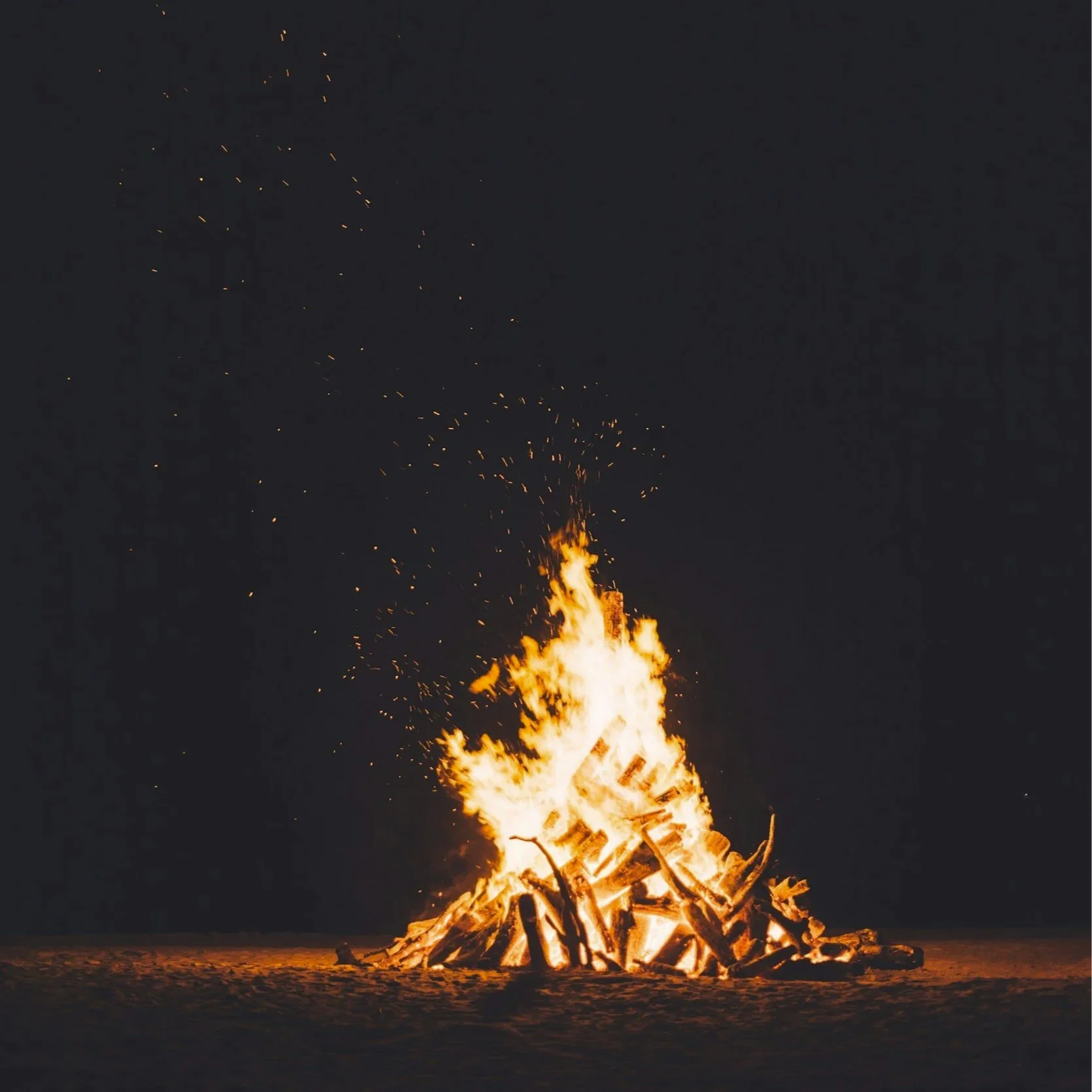 A large bonfire burning at night with bright flames and sparks flying into the dark sky.