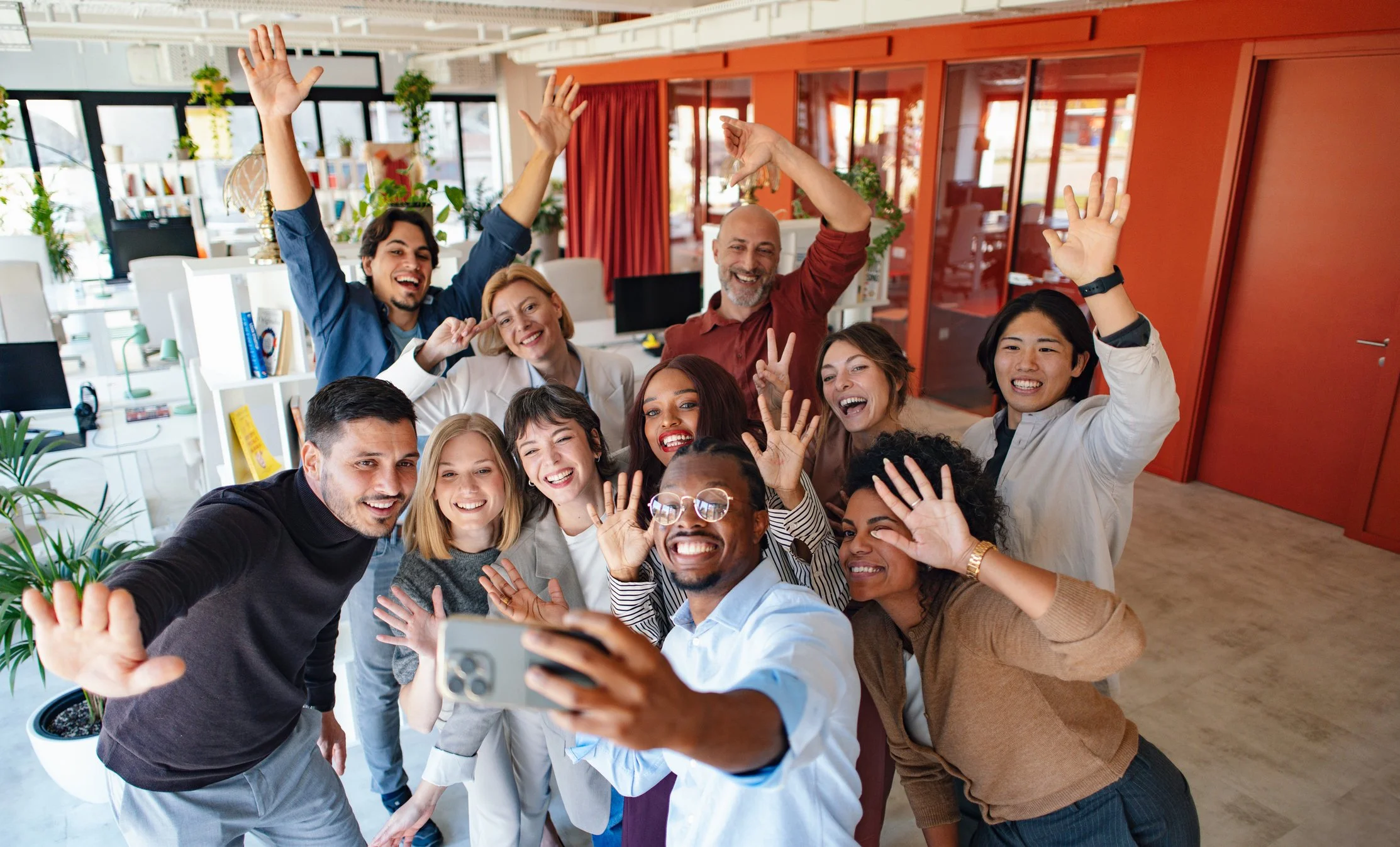 Group of diverse people taking a selfie together in an office, smiling and waving.
