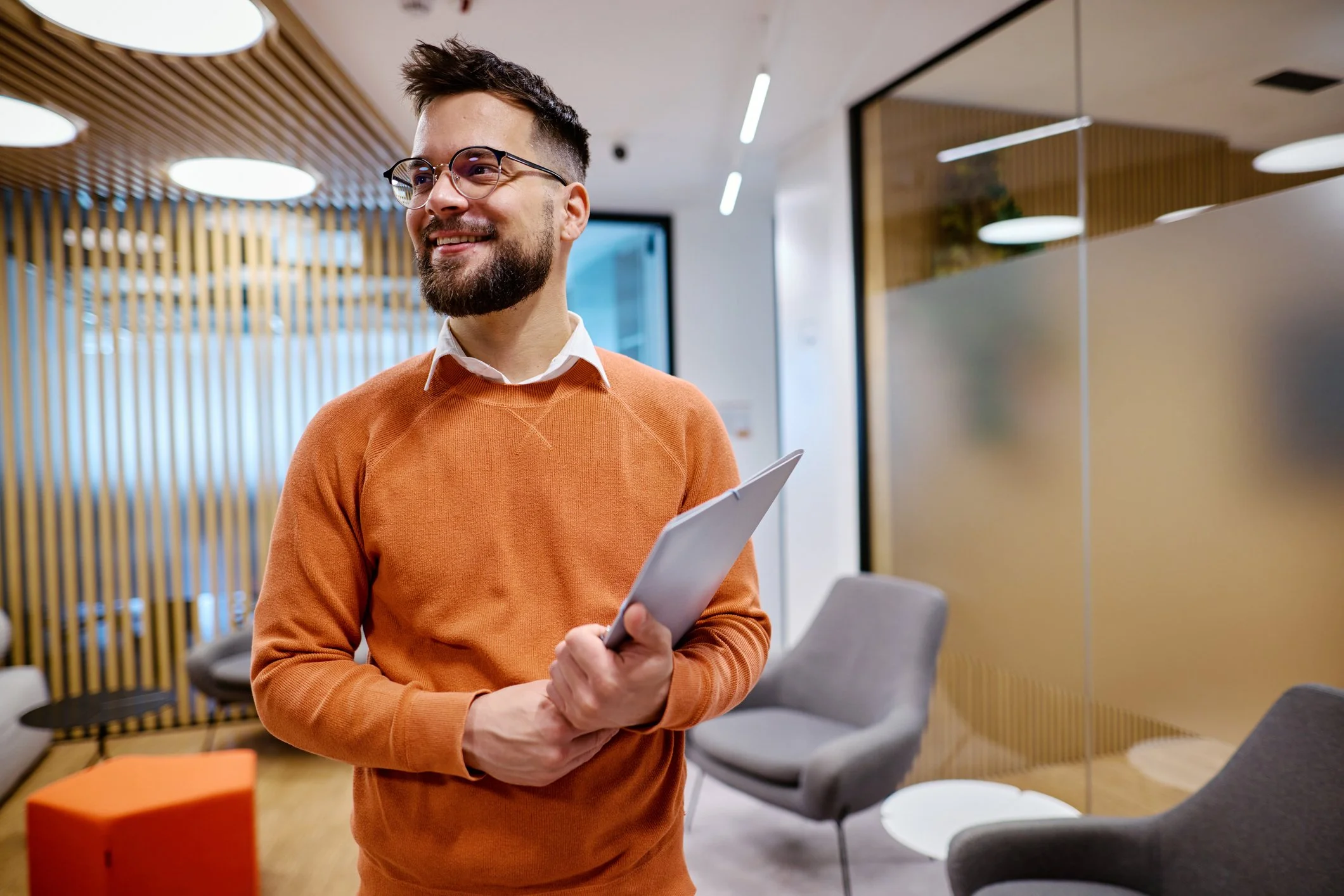 A smiling man with glasses and a beard, wearing an orange sweater and white shirt, standing in a modern office lobby holding a tablet.