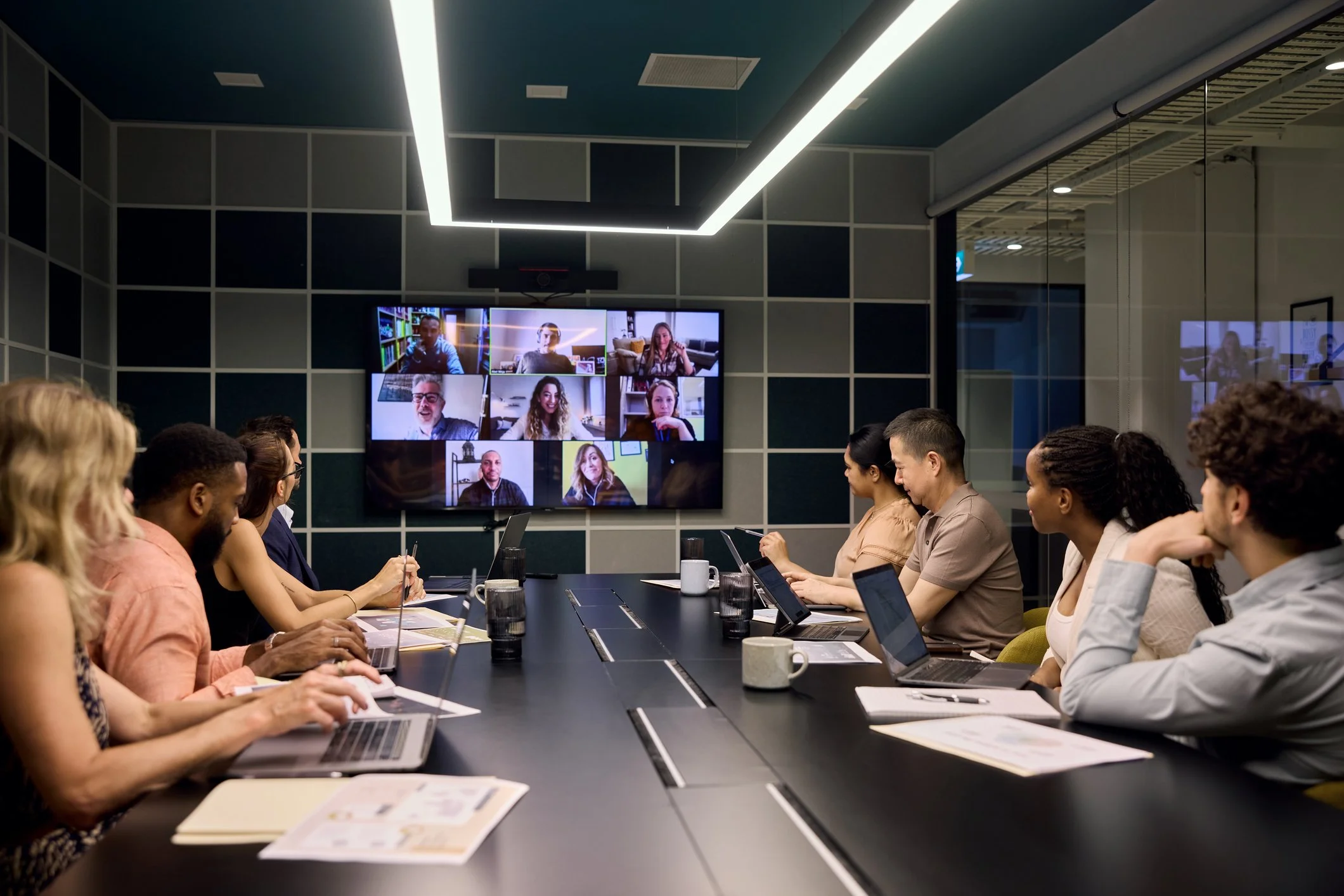 People sitting around a conference table, participating in a virtual meeting on a large screen, in a modern office conference room.