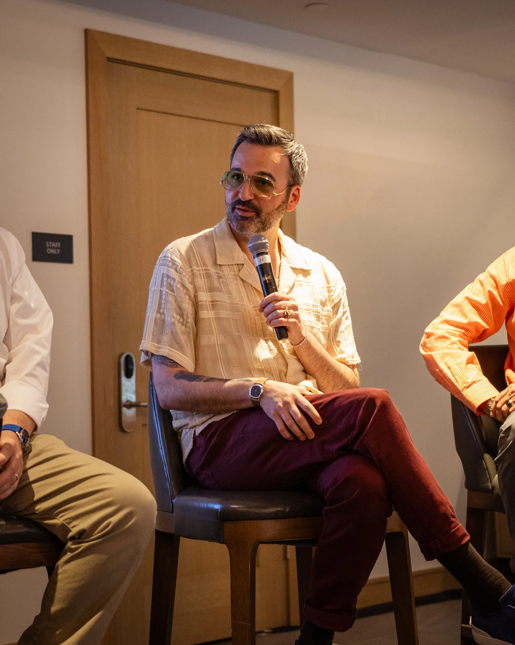 A man with glasses and a beard holding a microphone, sitting on a brown chair, wearing a beige shirt and maroon pants, participating in a panel discussion.