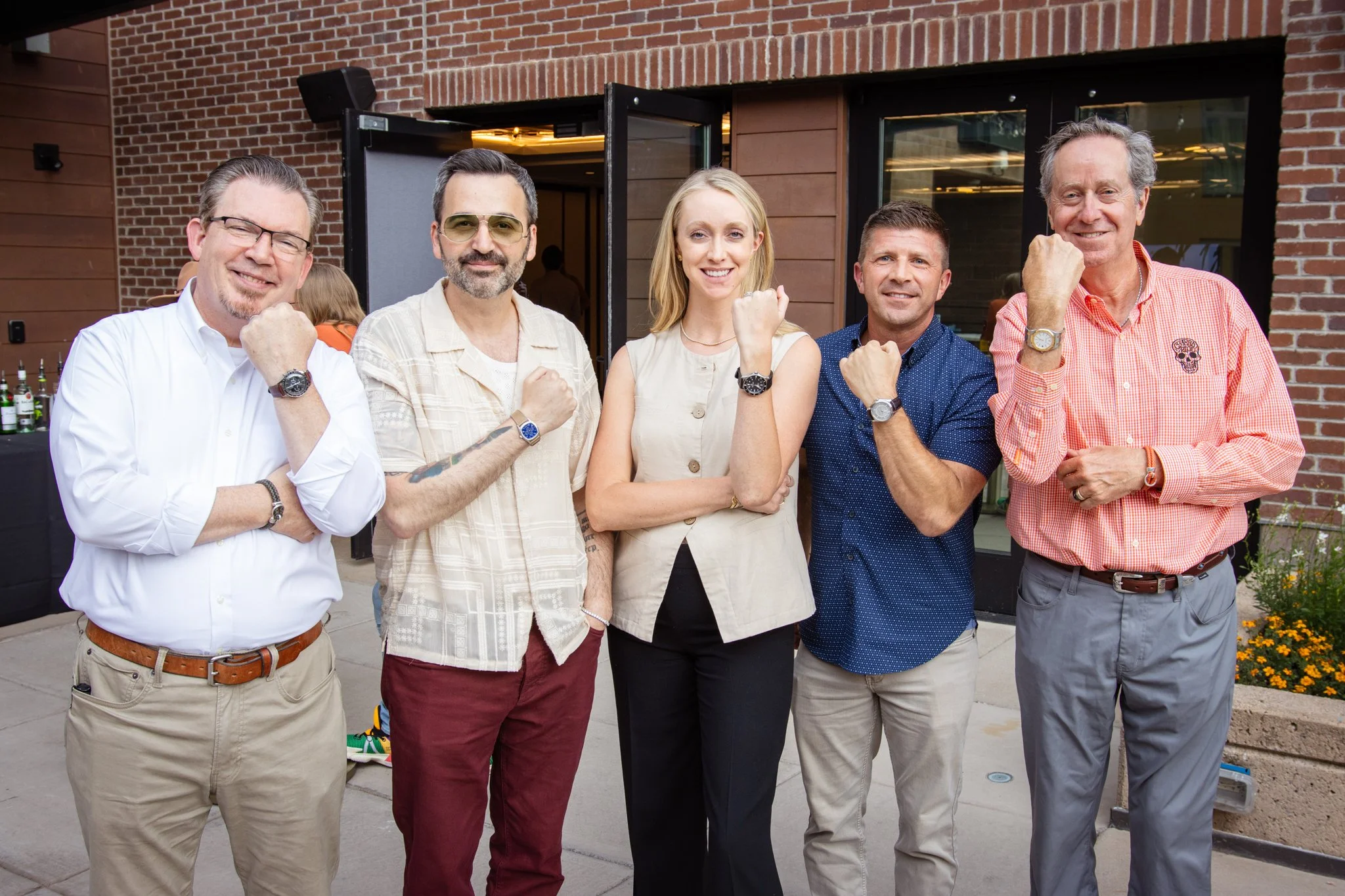 Group of six adults standing outside in front of a brick building, flexing their arms and smiling at the camera.