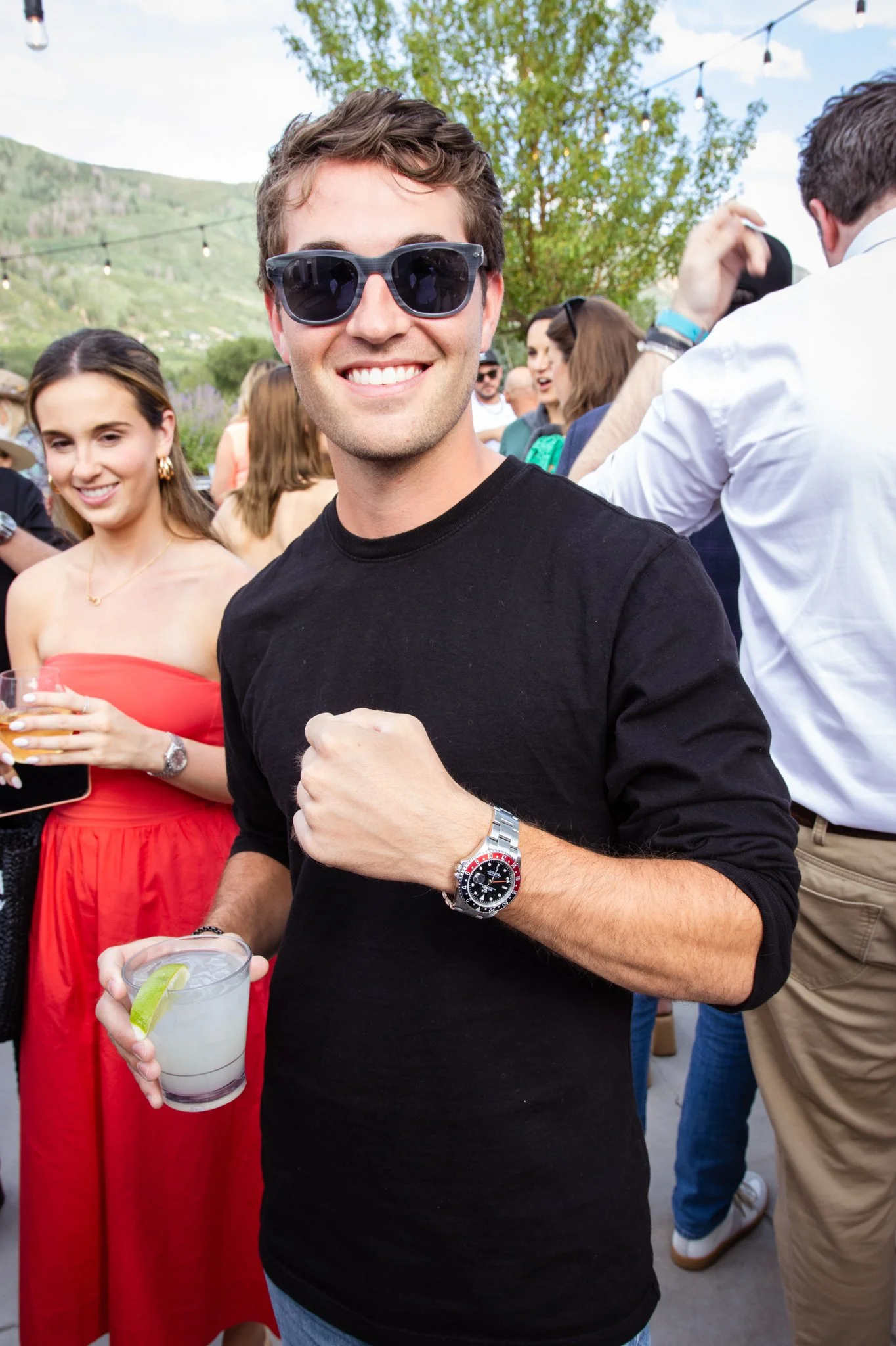 A young man wearing sunglasses, a black shirt, and a watch, holding a drink with a lime wedge at an outdoor party or gathering
