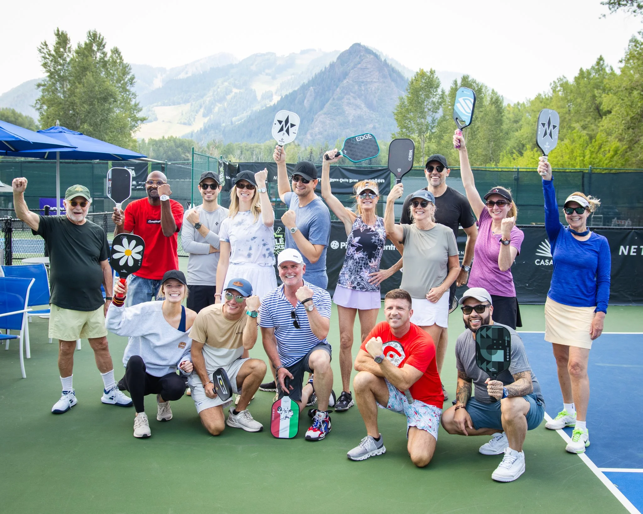A group of people on a tennis court celebrating after playing pickleball, holding paddles, and making fists with smiles, with trees and mountains in the background.