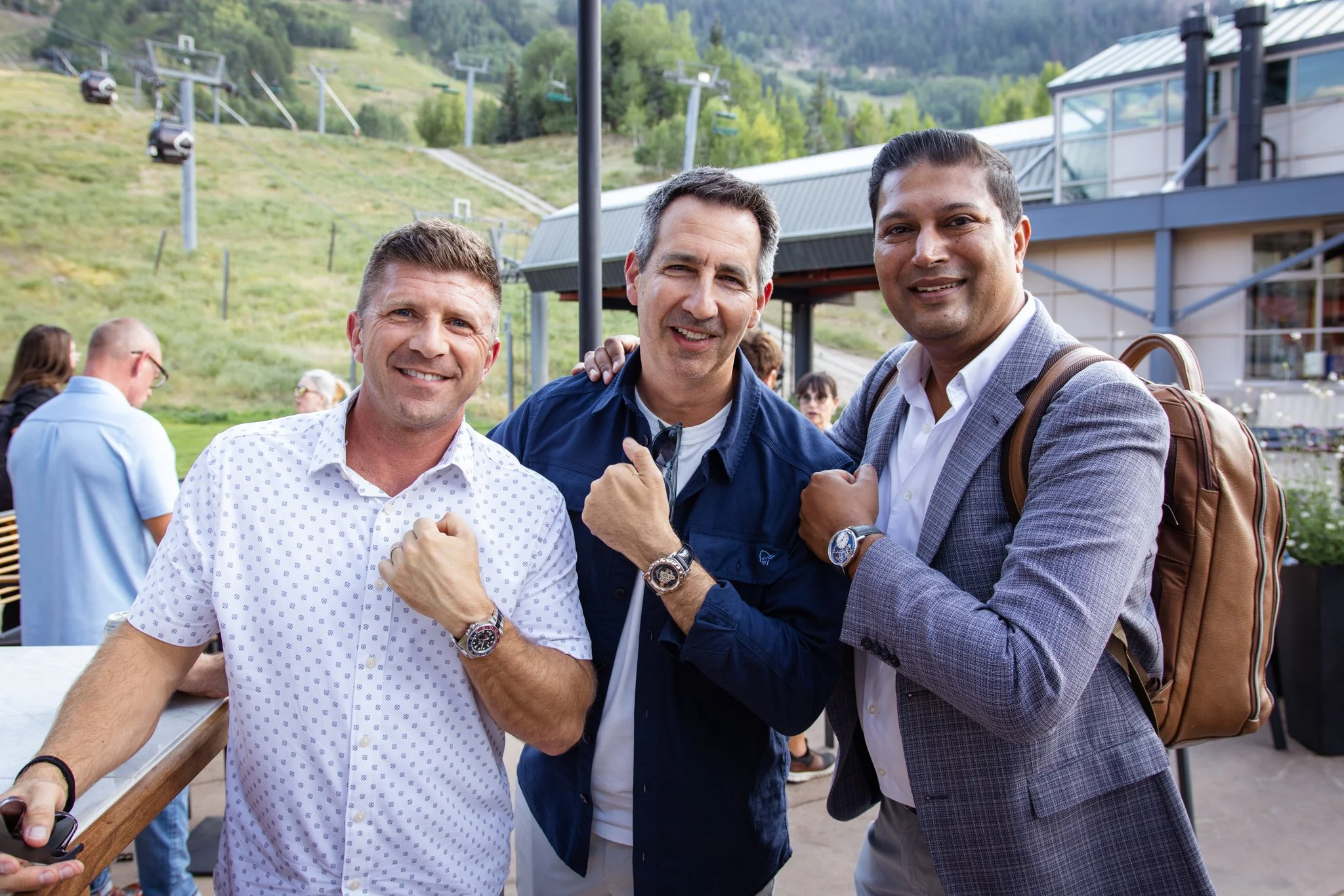 Three men smiling at an outdoor social gathering, standing close together with fists raised, wearing watches, with a mountainous landscape and ski lift chairs in the background.